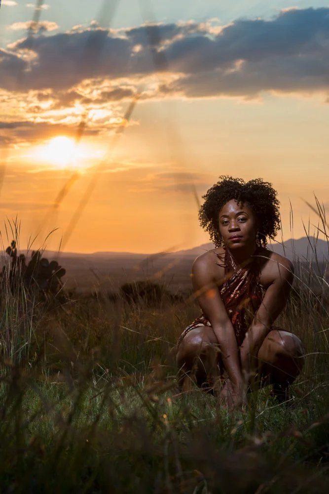 A woman with curly hair squatting in a grassy field during sunset, with the sun partially obscured by clouds in the background.