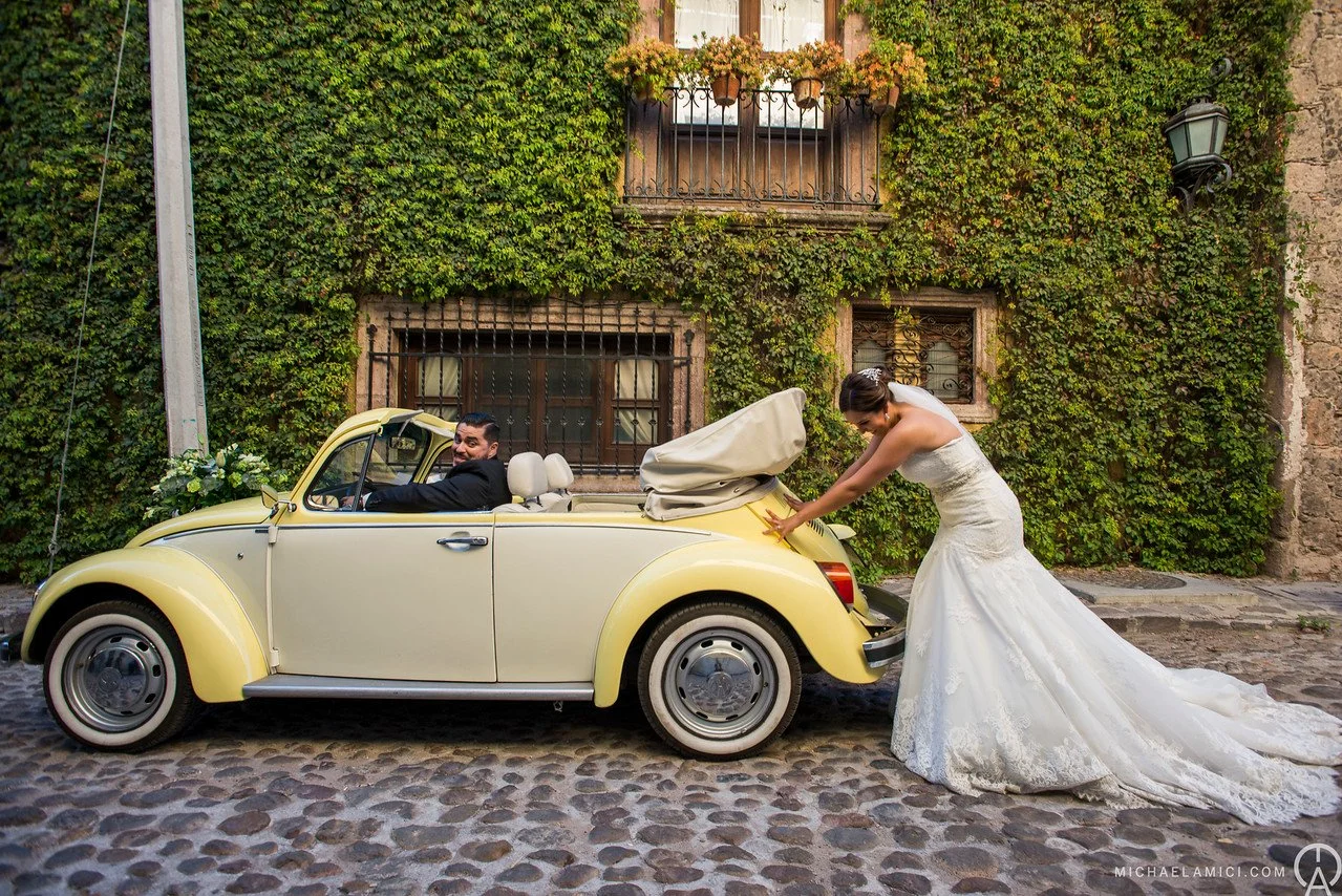 A bride in a white wedding gown leaning into the trunk of a vintage yellow Volkswagen Beetle, with a groom sitting inside, smiling. The scene is set on a cobblestone street with a green ivy-covered wall in the background.