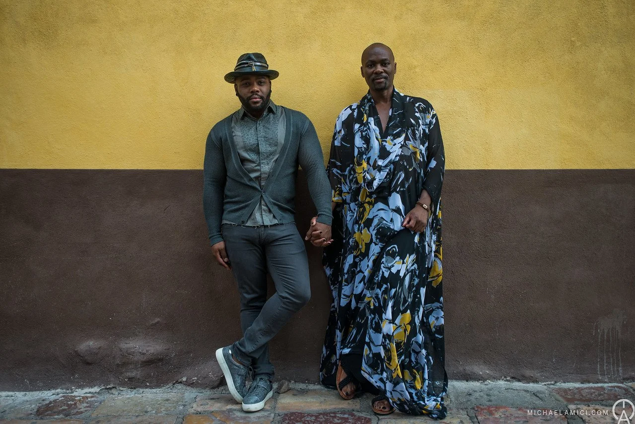 Two men standing against a yellow and brown wall, holding hands and looking at the camera. One man is dressed in modern casual clothing with a hat, and the other is wearing a long, floral-patterned dress.