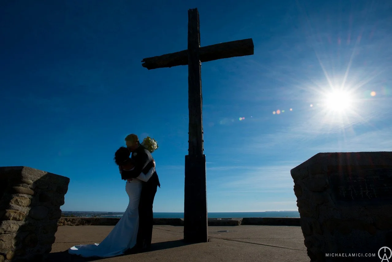 Silhouette of a newlywed couple embracing and kissing under a large wooden cross during sunset, with a clear sky and ocean in the background.