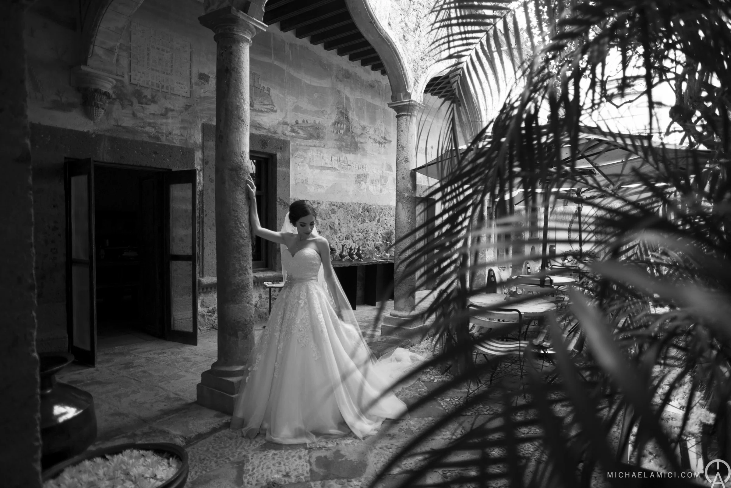 A bride in a wedding dress standing inside a historic stone building with columns, surrounded by tropical plants, with a mural on the wall behind her.