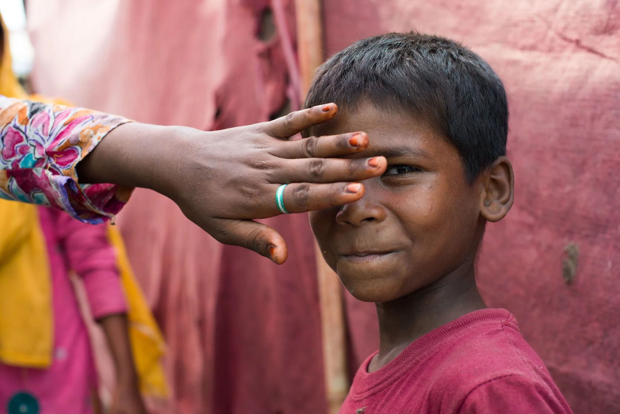 A young boy with short black hair and a reddish shirt has his forehead gently touched by an adult's hand with painted fingernails. The background features a pinkish wall.