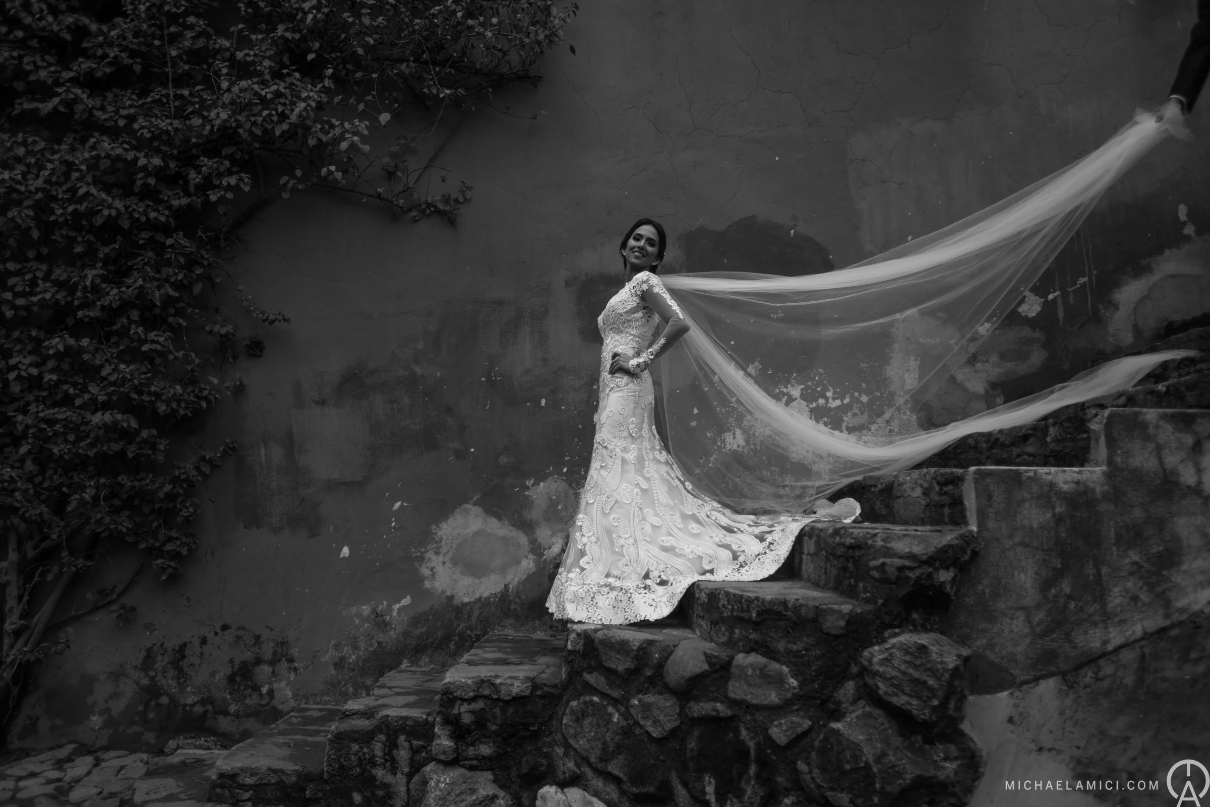 Woman in an elegant wedding dress standing on stone steps against a textured wall, with a long veil flowing behind her.