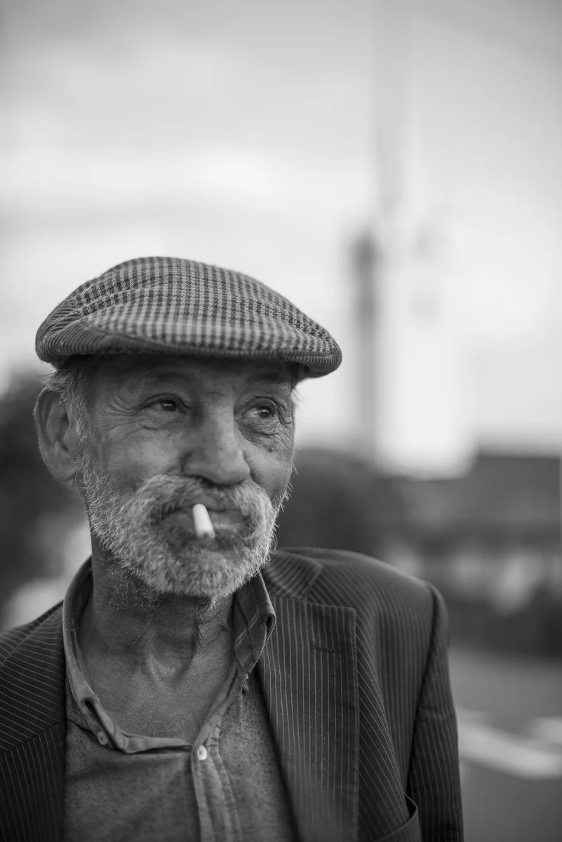 Black and white photo of an elderly man with a beard and mustache, wearing a flat cap and a striped blazer, with a cigarette in his mouth, outdoors.