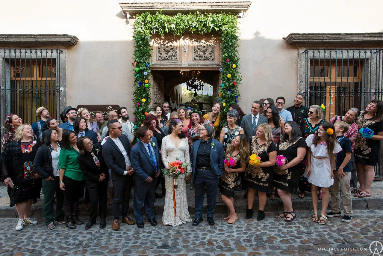 A diverse group of people celebrating a wedding outside a building with a decorative entrance, some holding colorful flowers, others smiling and looking at the bride and groom, who are standing in the front center.