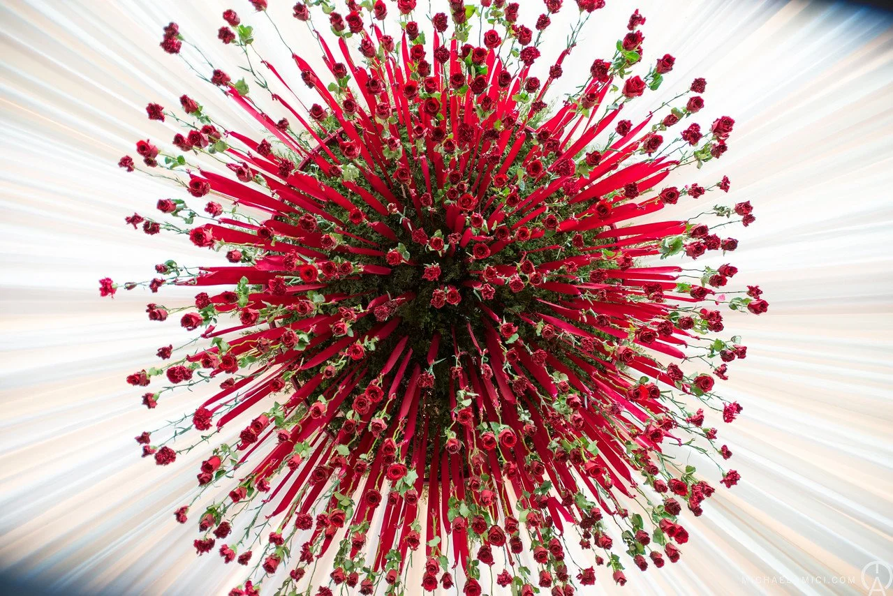 A floral arrangement with many small red roses and long red ribbons radiating outward in a spherical shape.