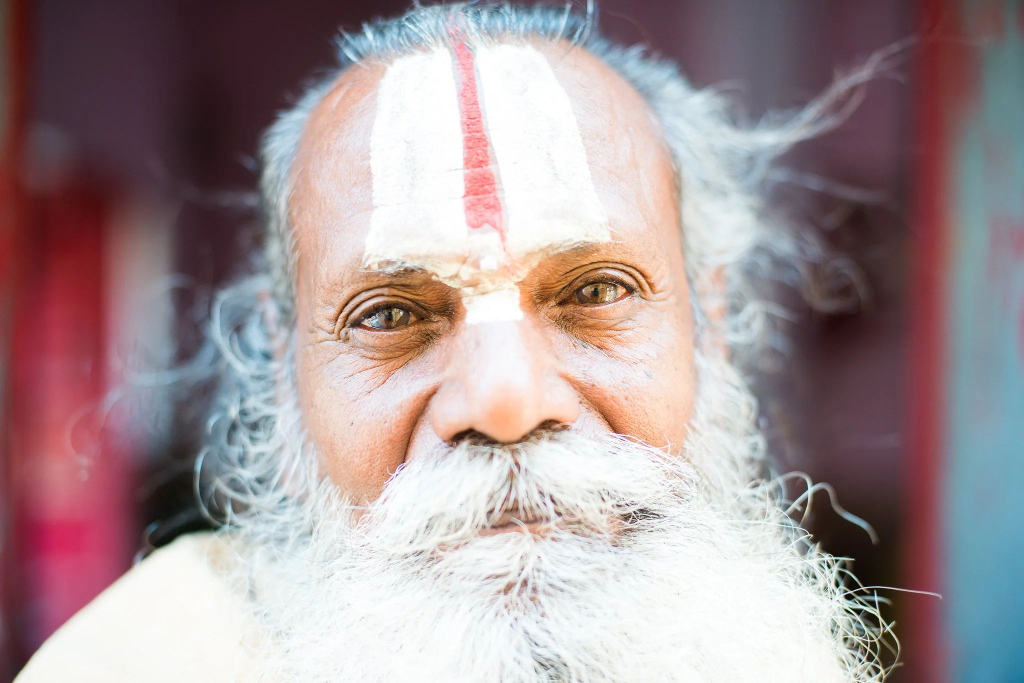 Close-up of an elderly man with a long white beard, white hair, and a white and red mark on his forehead, smiling softly.