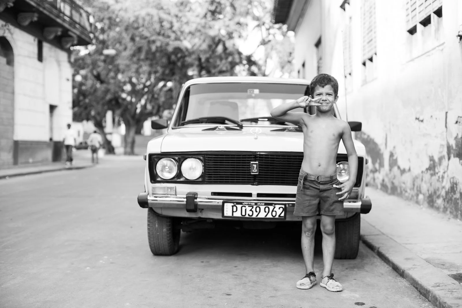A shirtless young boy in shorts and sneakers stands on a street in front of a vintage car, making a peace sign near his eye, with trees and buildings in the background.