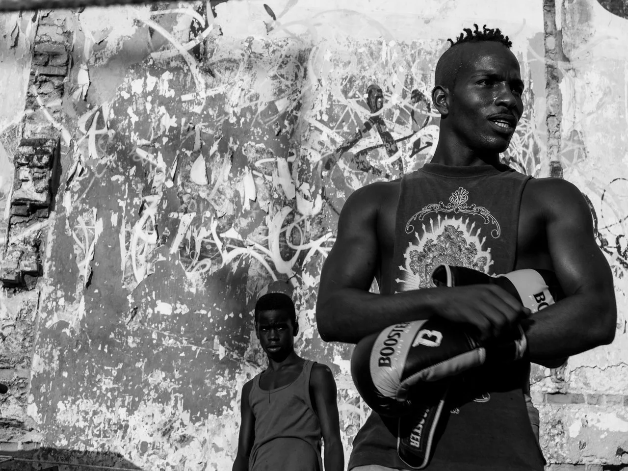 Two young men stand in front of a wall with graffiti in an urban setting. The man in the foreground is holding a boxing glove, wearing a sleeveless shirt, and looking to the side. The second man is further back, wearing a tank top, and looking direct