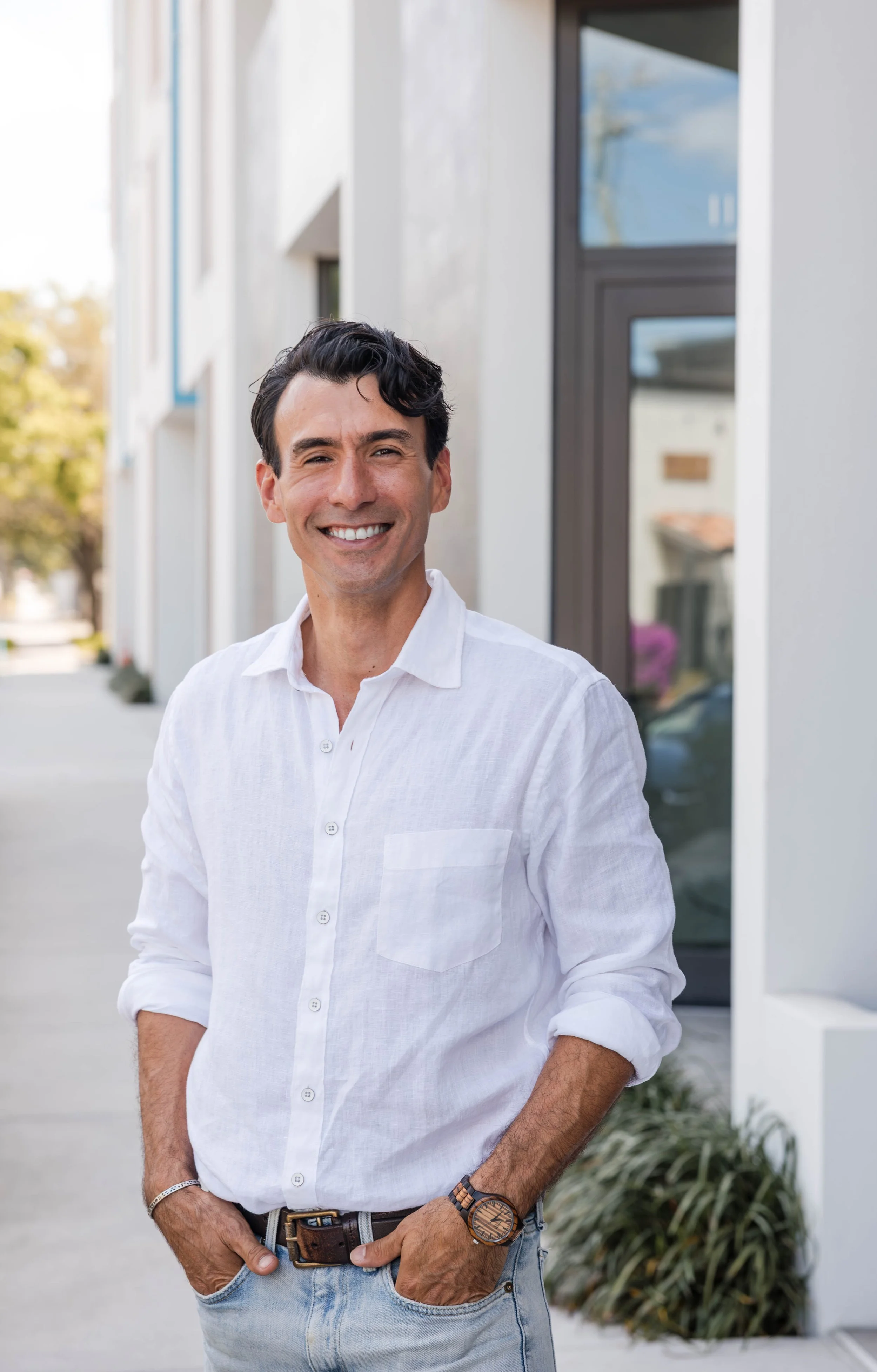 A man standing outdoors smiling, wearing a white button-down shirt with rolled-up sleeves, light blue jeans, and a wooden wristwatch.