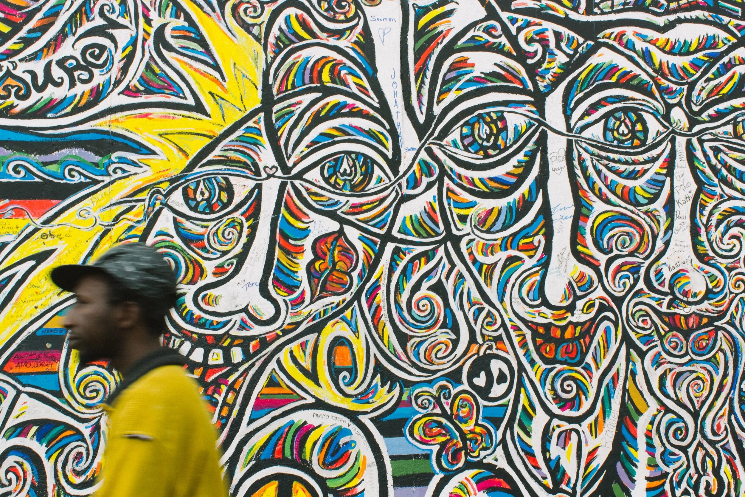 A colorful street art mural featuring a large face with swirling patterns, butterflies, and abstract designs, with a person in a yellow jacket and black cap walking past in the foreground.