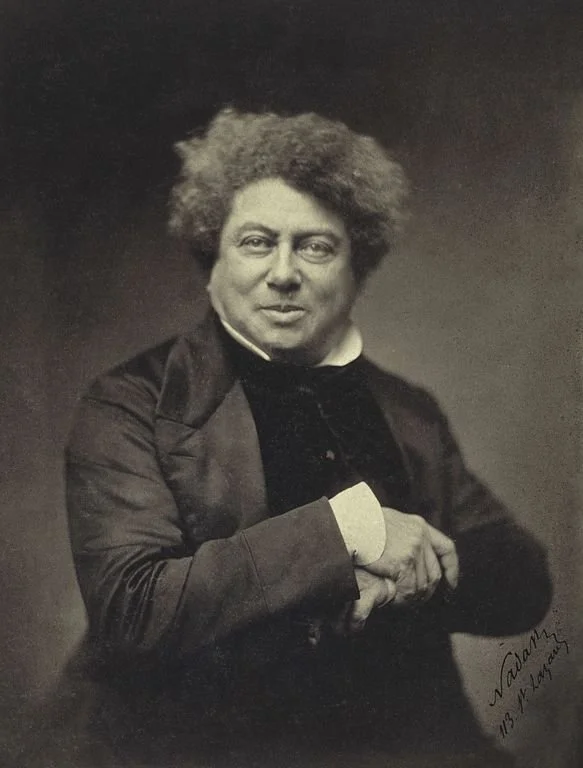 Alexandre Dumas B&W photo taken in 1855. A middle-aged man with frizzy hair in a suit with collar