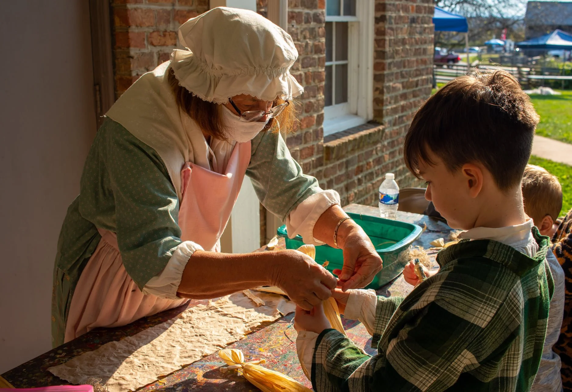 Woman in historical attire assists children with crafts outdoors.