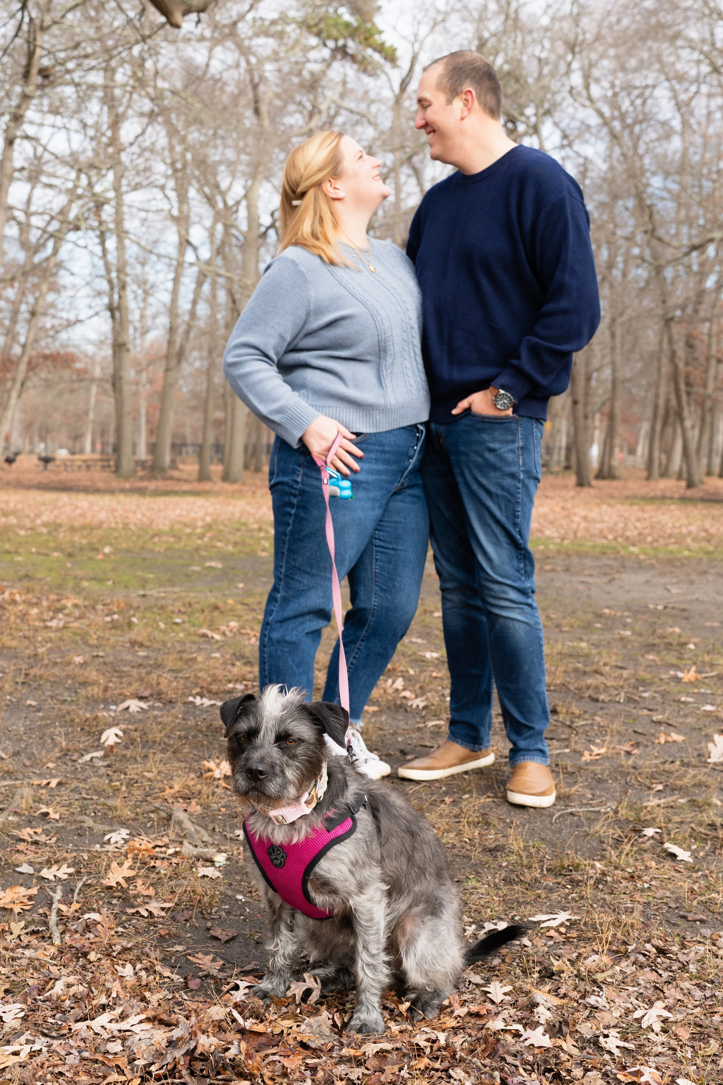 A couple smiling and facing each other in a park with fallen leaves and trees in the background, standing with a dog on a leash.