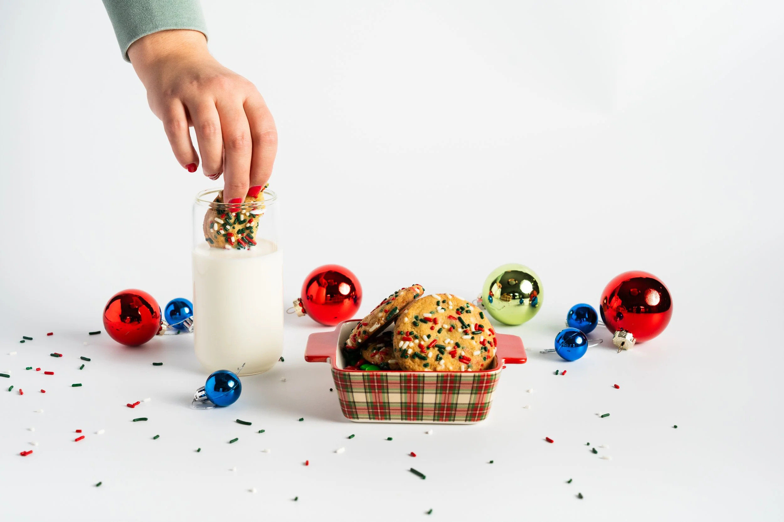 Decorative Christmas cookies with sprinkles in a tart pan, a hand dunking a cookie into a glass of milk, surrounded by colorful Christmas ornaments and confetti on a white background.