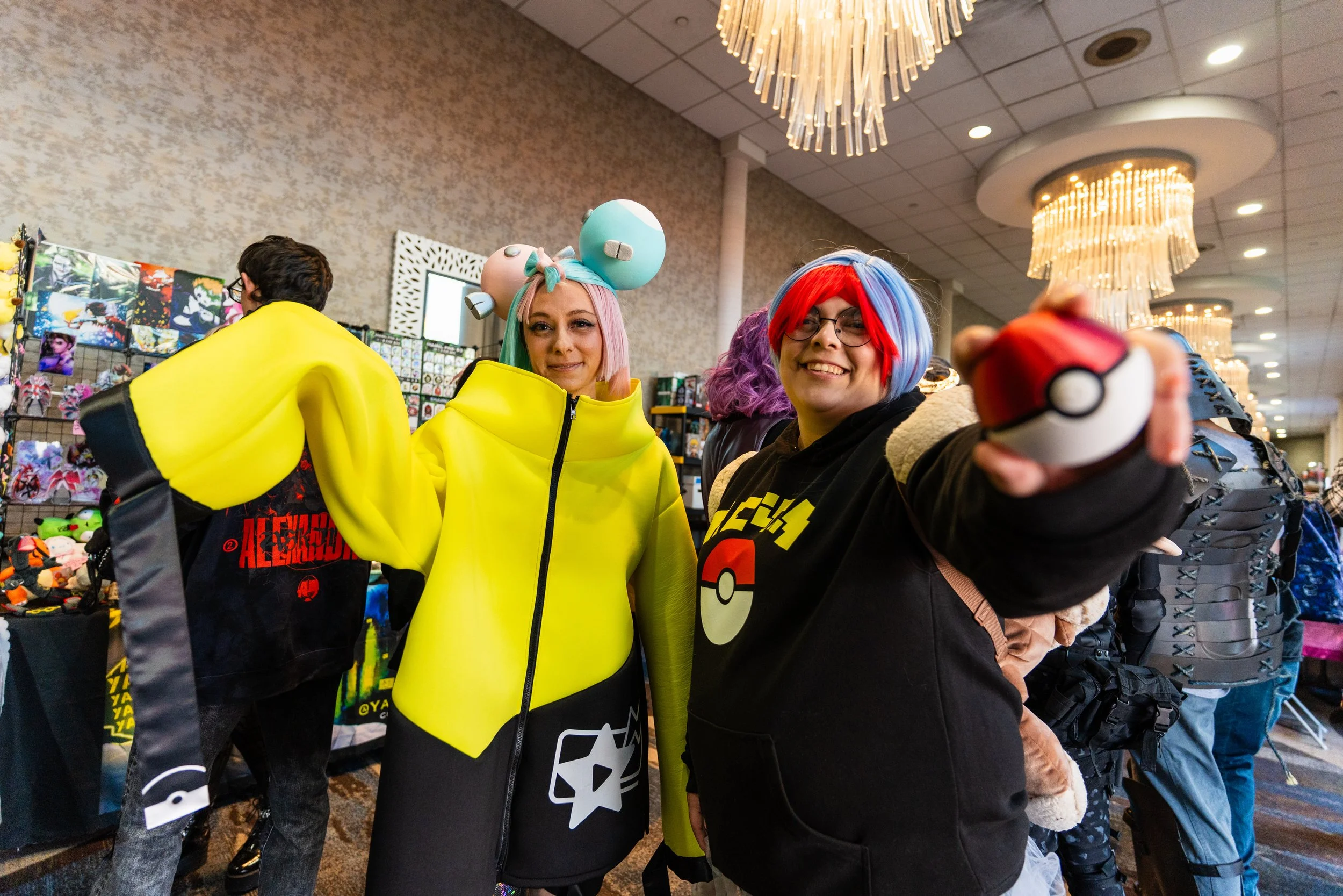 Two people dressed as anime characters at a convention, one with pink hair and pastel-colored accessories, the other with multicolored hair and glasses, holding a Pokéball.