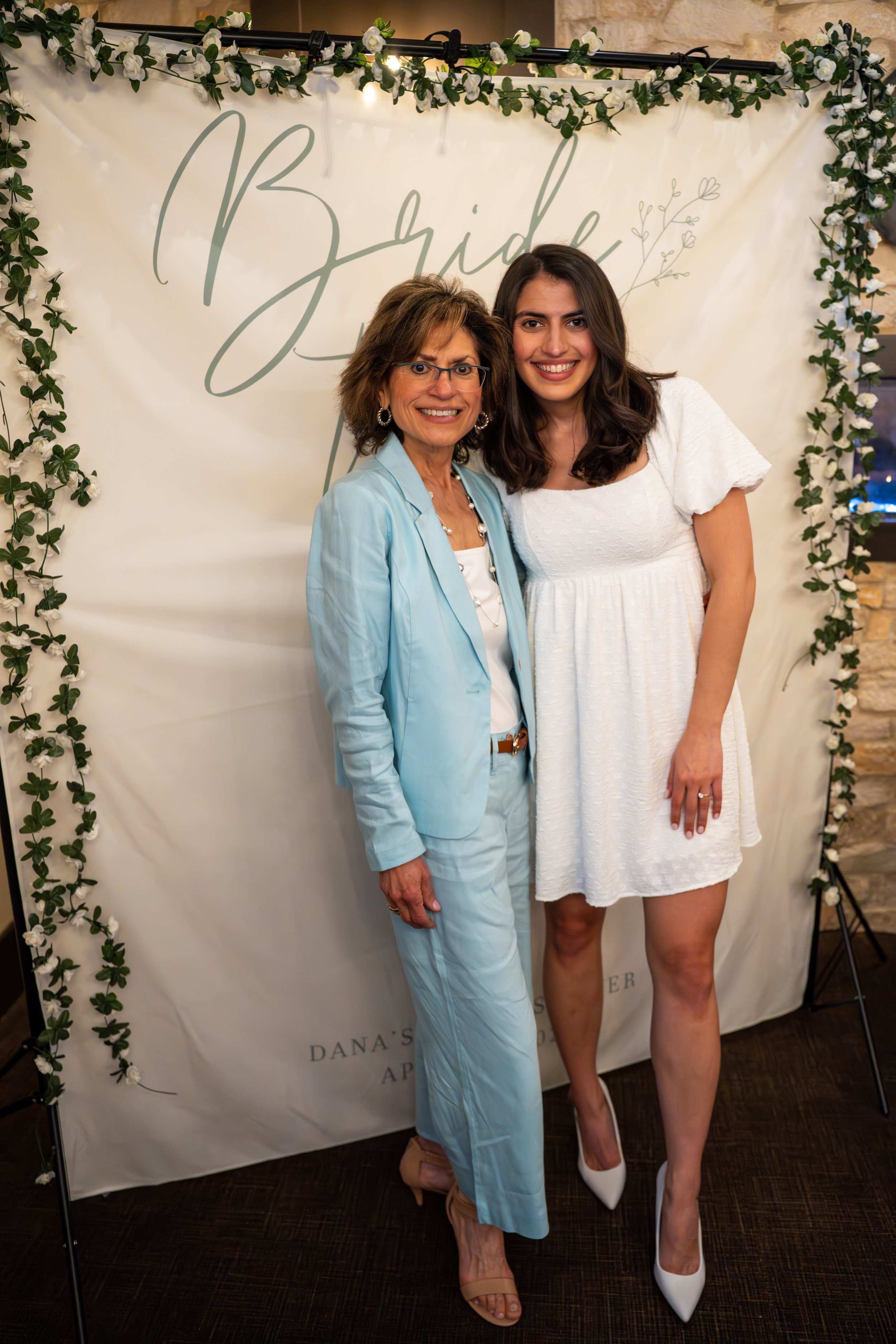 Two women standing together and smiling in front of a decorative backdrop that says 'Bride'. The older woman is wearing a light blue suit with a white top and beige heels. The younger woman is dressed in a white dress with white heels. The backdrop is decorated with greenery and small white flowers.
