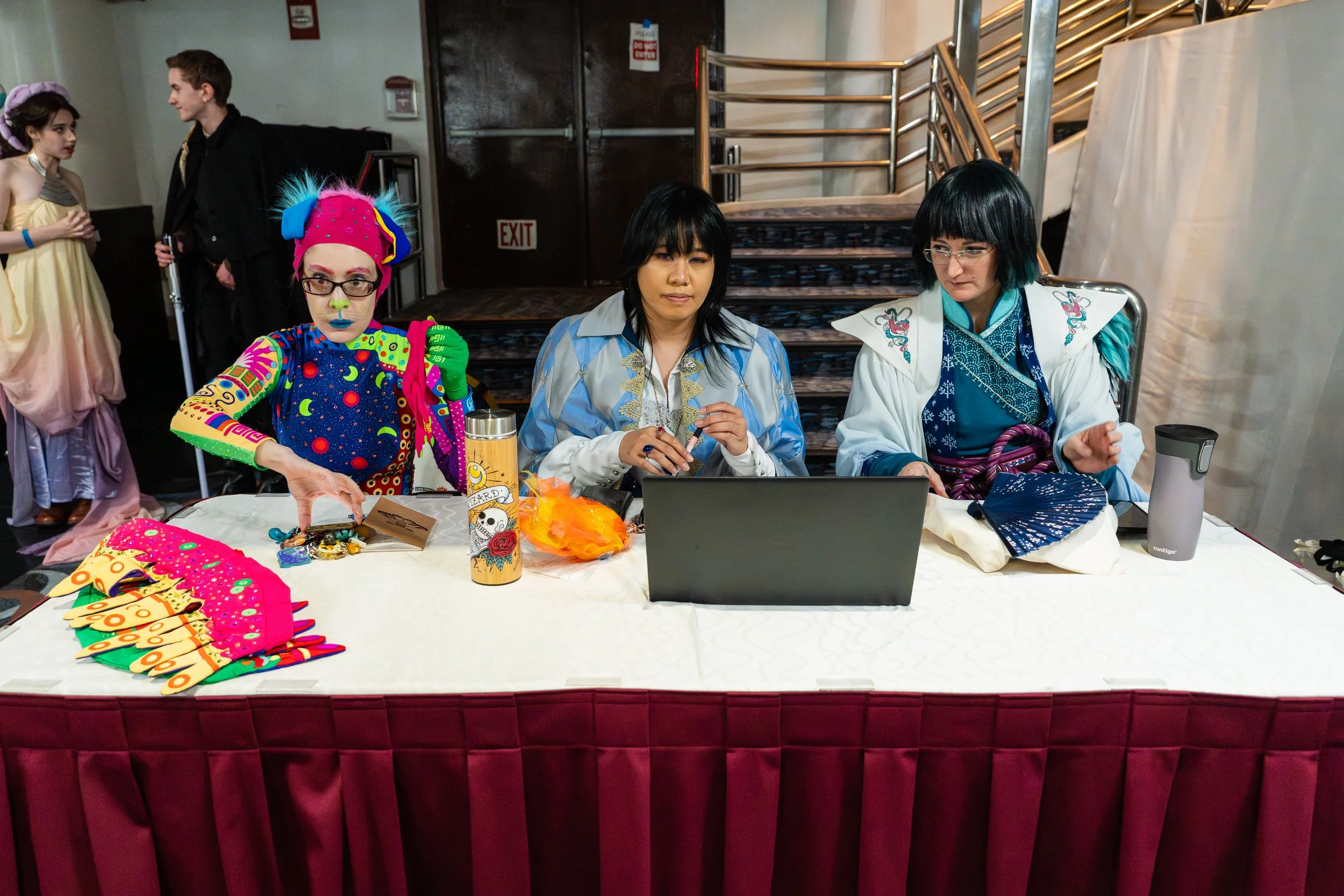 Three people dressed in colorful costumes sitting at a table, with two kids in costumes in the background.