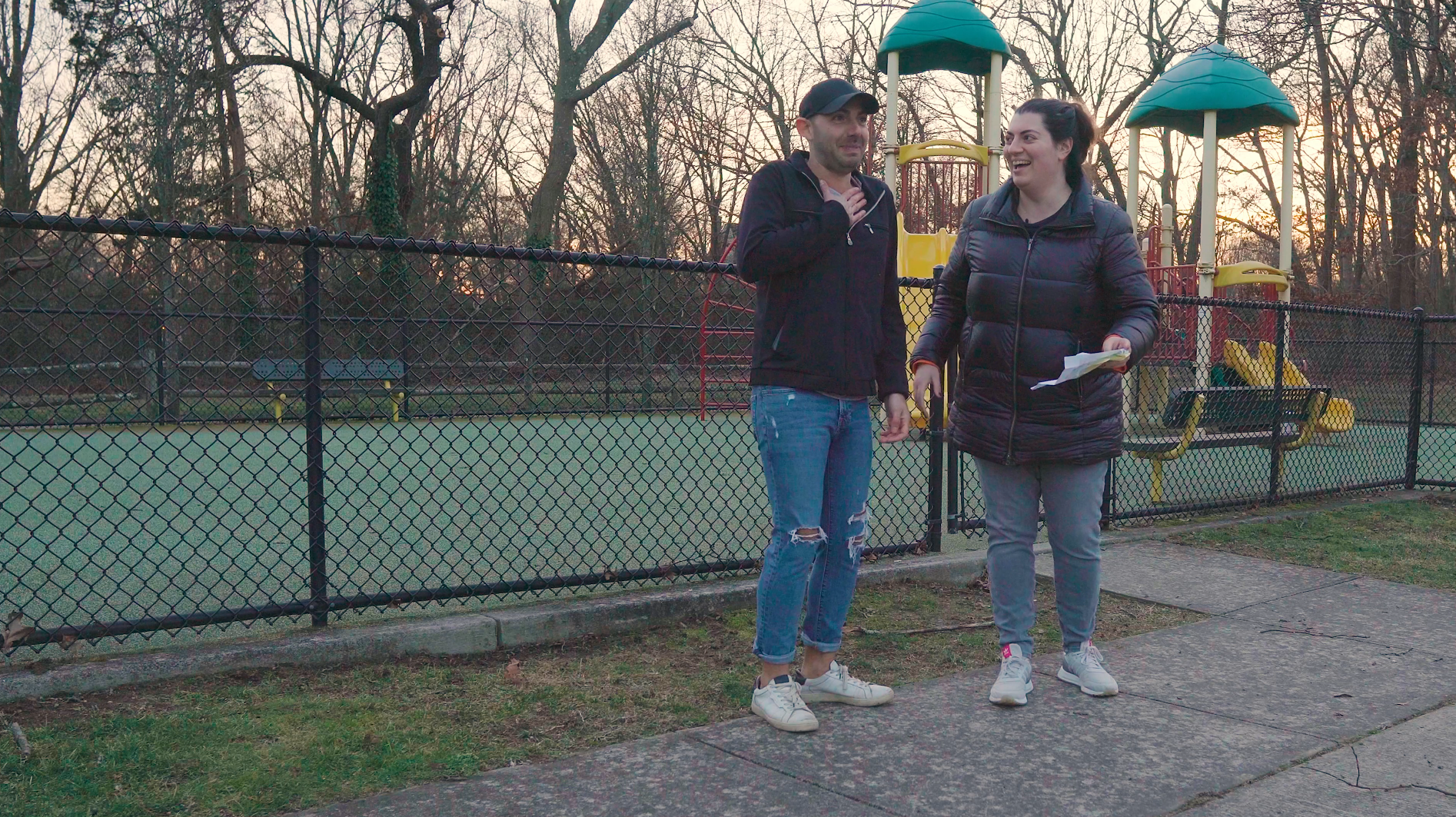 Two people, a man and a woman, are standing and talking next to a playground with a fenced tennis court in the background. The man is wearing a black jacket and jeans with ripped knees, and the woman is wearing a black puffer jacket and gray pants. They appear to be engaged in a conversation.