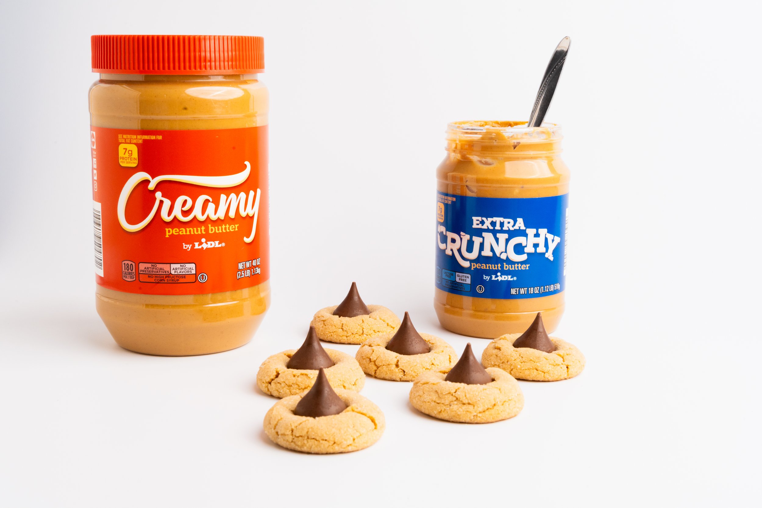 A jar of creamy peanut butter, a jar of extra crunchy peanut butter with a spoon inside, and six cookies with chocolate peanut butter peaks on top on a white background.