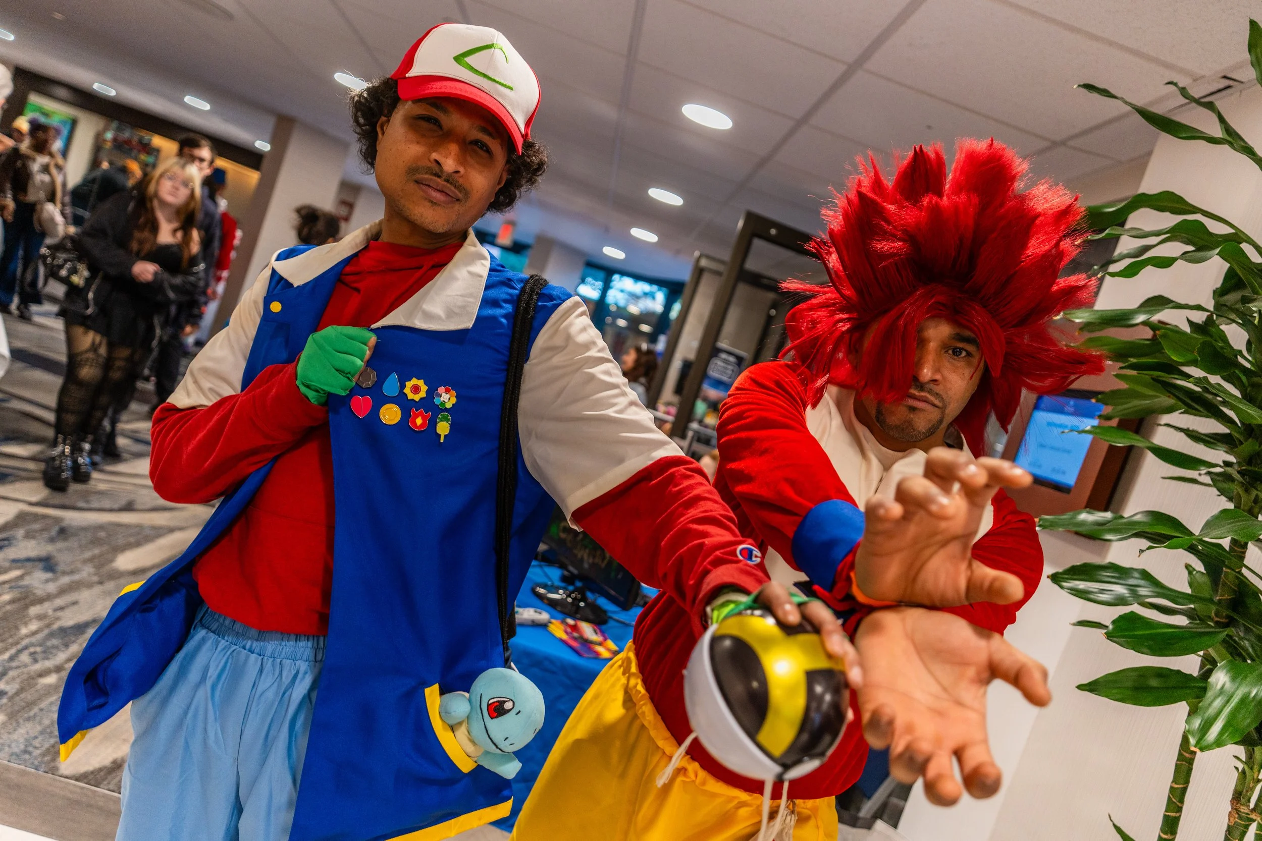 Two men dressed as Pokémon characters, one as Ash Ketchum with a red and white hat, blue vest, and green gloves, and the other as Jessie with a large red wig and red outfit, posing with their hands extended towards the camera at a convention or event