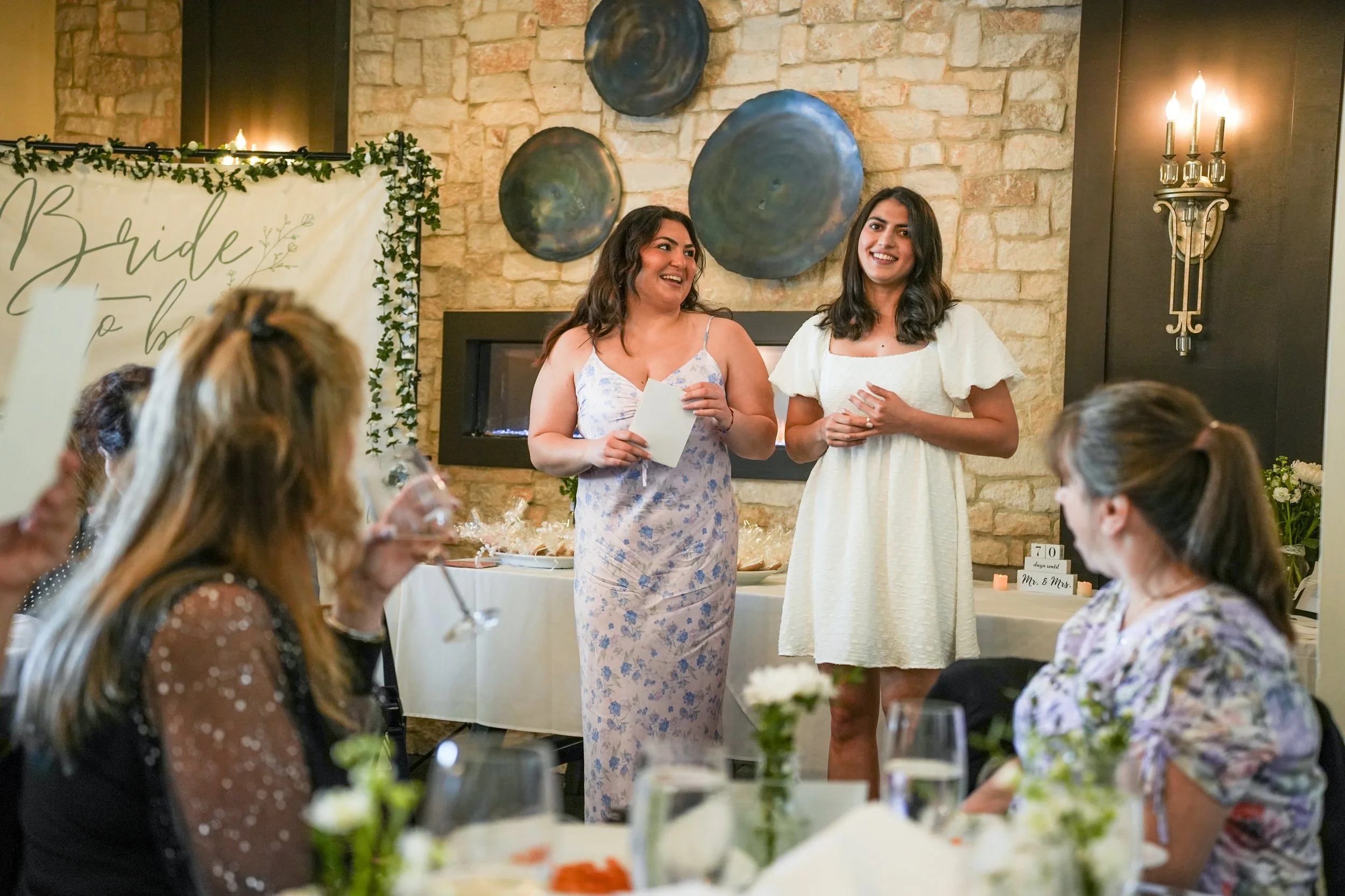 Two women standing and smiling at a wedding reception with seated guests, a decorative sign, and wall art in the background.
