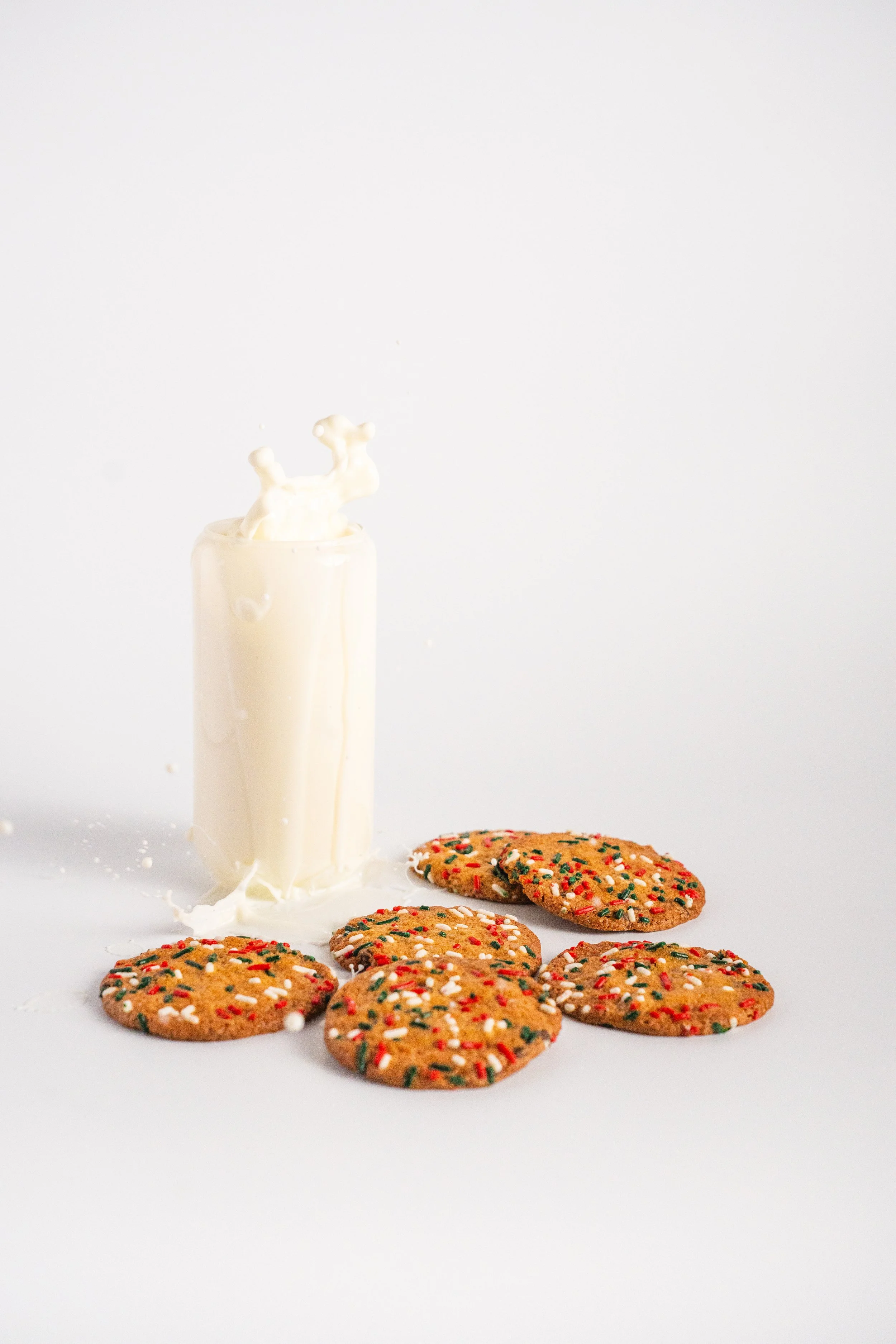 A glass of milk with a cookie on a white background. The cookie is decorated with red, green, and white sprinkles.