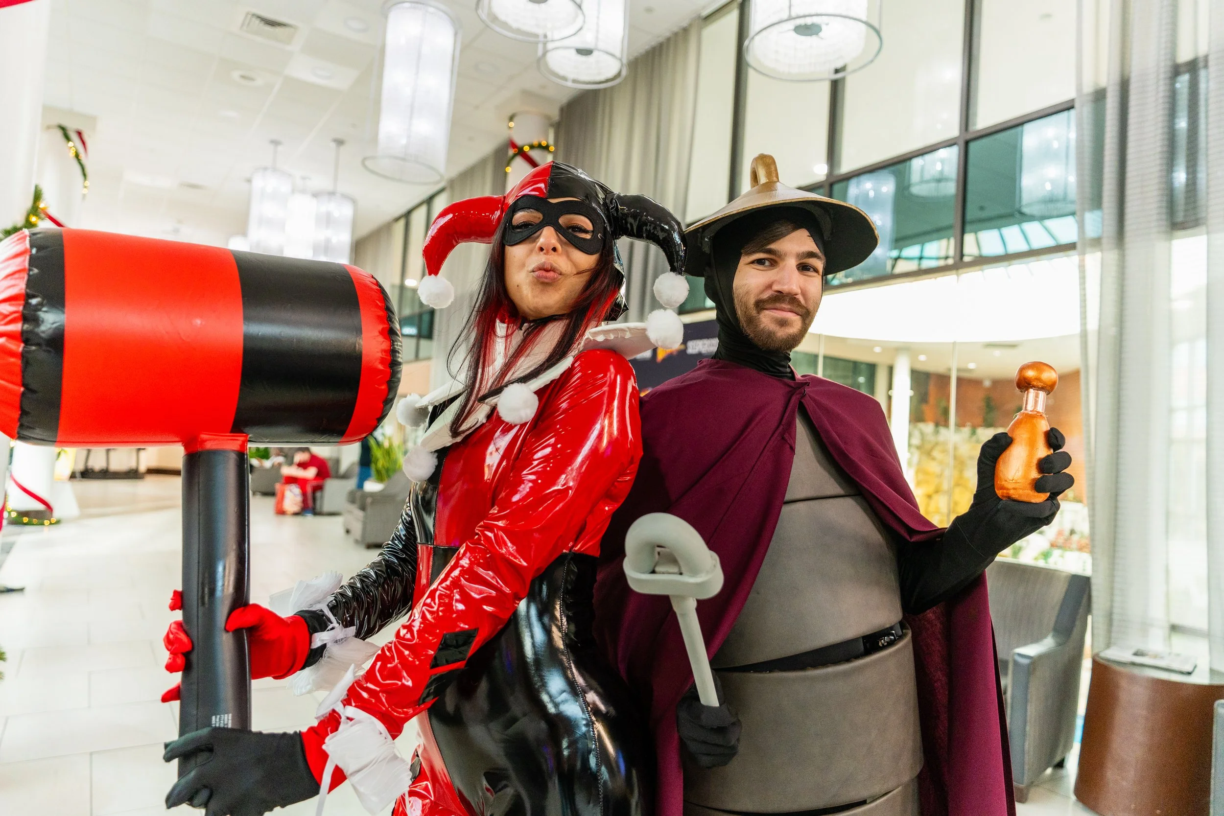 Two people dressed in costumes, one as Harley Quinn and the other as a wizard, standing back to back in a modern indoor setting.