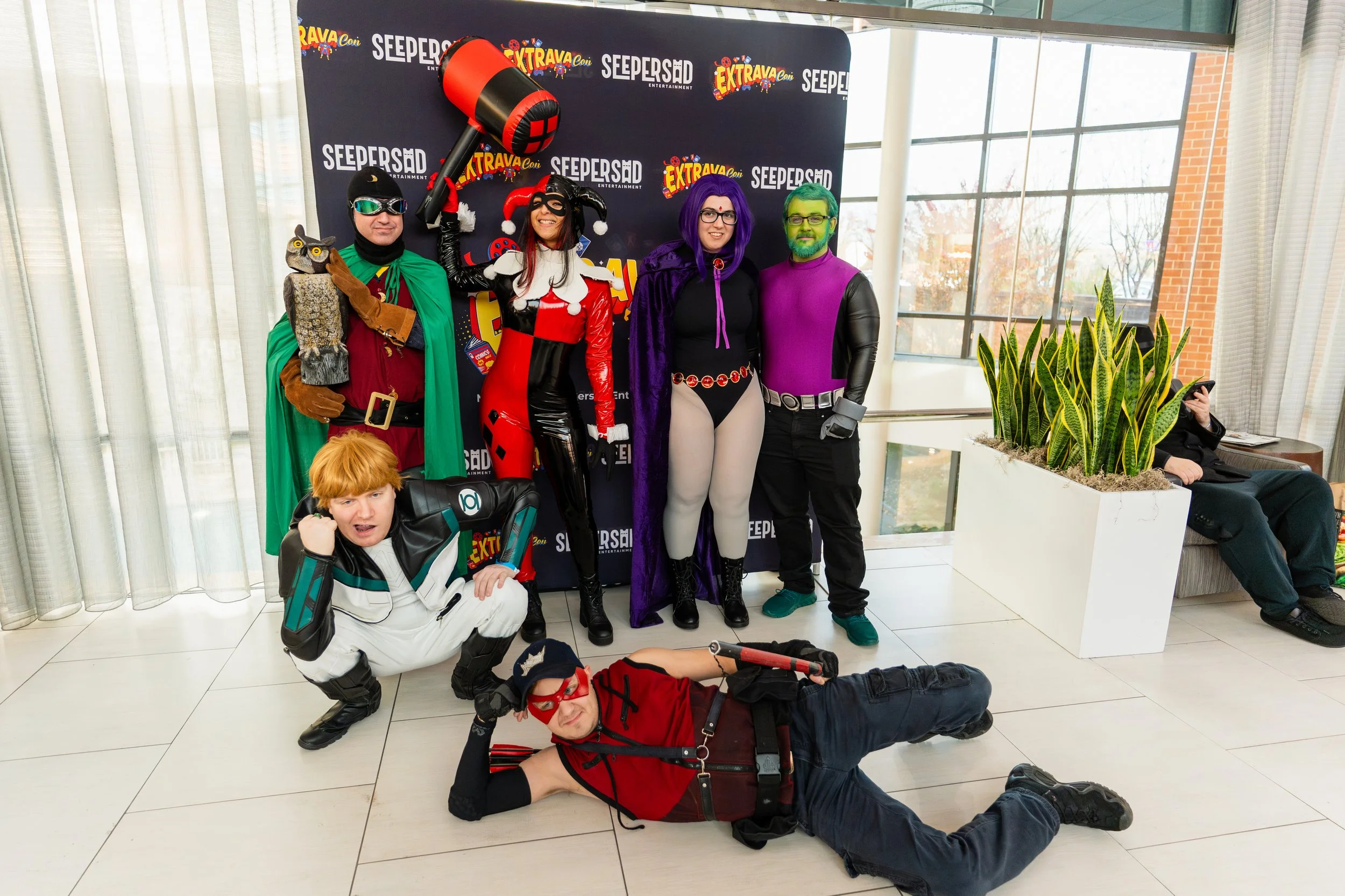 Group of eight people dressed in various cosplay costumes posing at a convention with a backdrop featuring logos and a large window behind them.