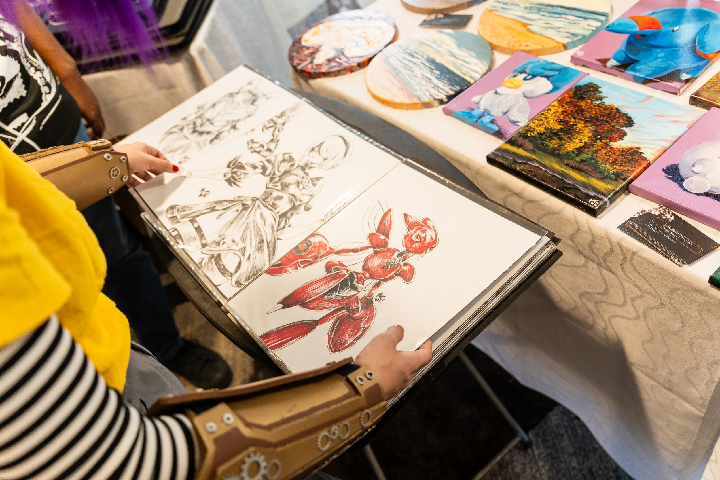 Person viewing sketches and various colorful paintings on a table at an art exhibit.