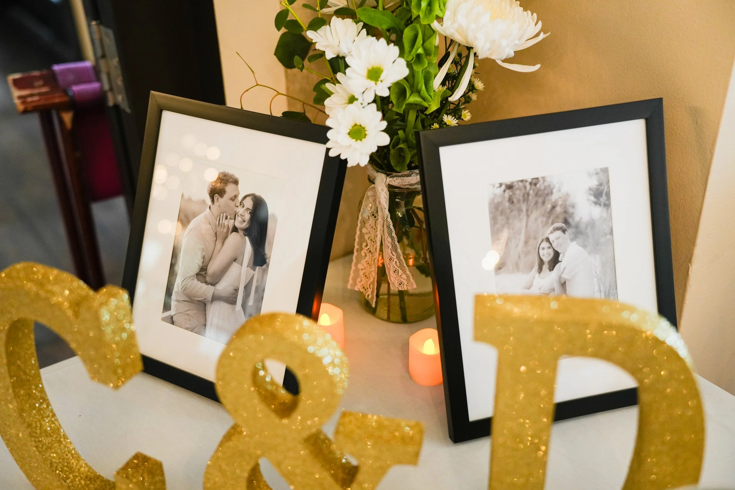 Two framed black-and-white wedding photographs displayed on a table with a bouquet of white flowers in a vase and some lit candles. Decorative gold ampersand symbol in the foreground.