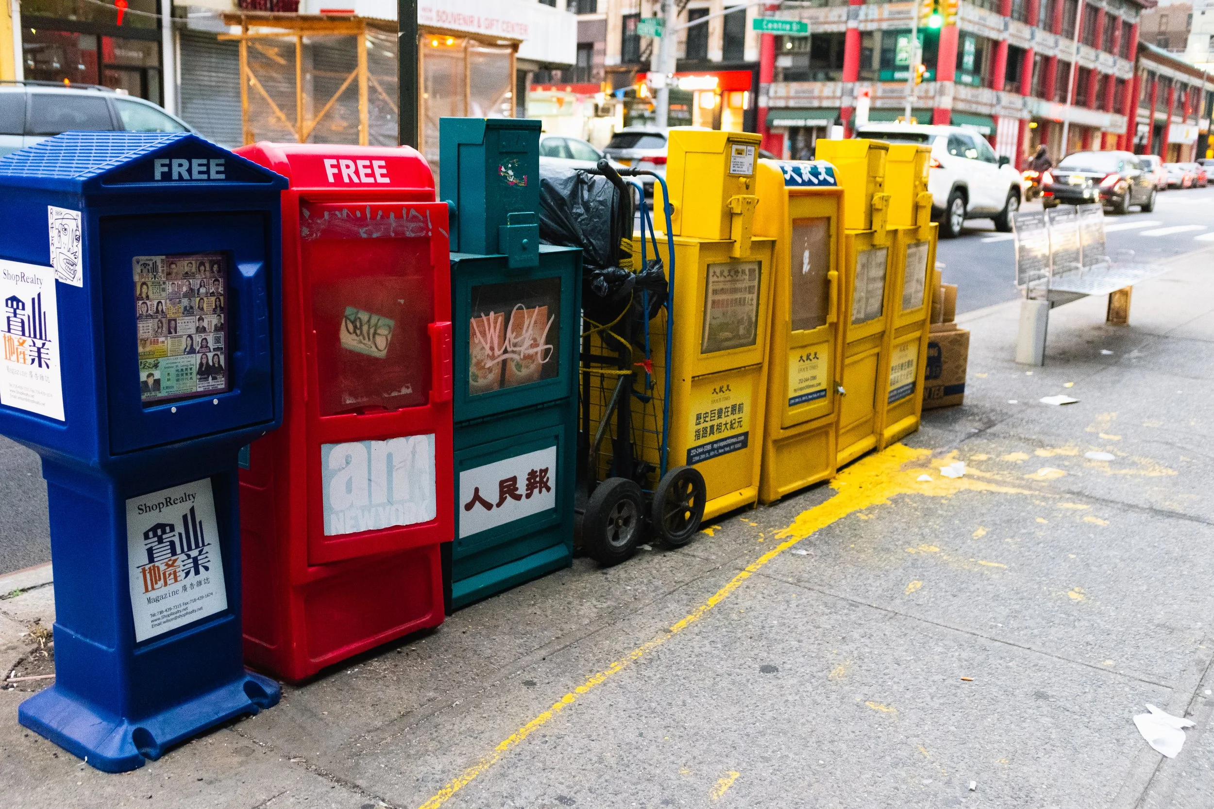 A row of colorful newspaper vending machines on a city sidewalk, including a blue, red, green, and yellow ones, with cars and buildings in the background.