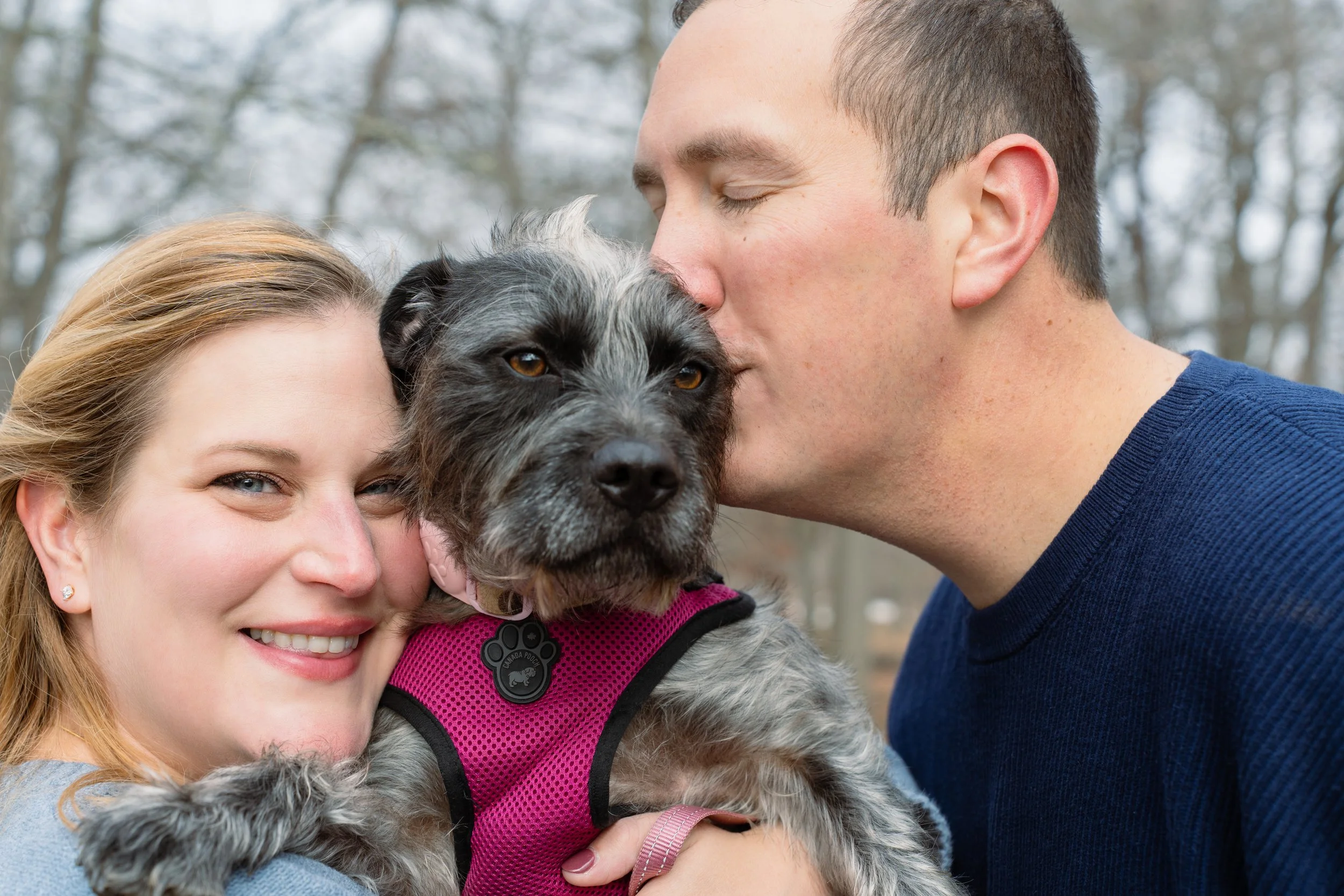 A smiling woman and a man kissing a dog in an outdoor park with bare trees.