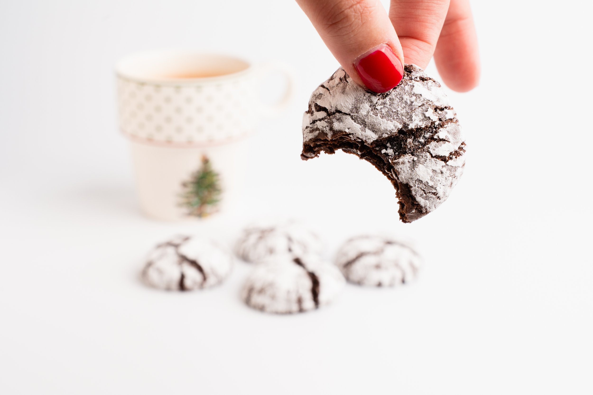 Hand with red nail polish holding a dusted chocolate crinkle cookie over a white surface with additional cookies and a festive cup in the background.