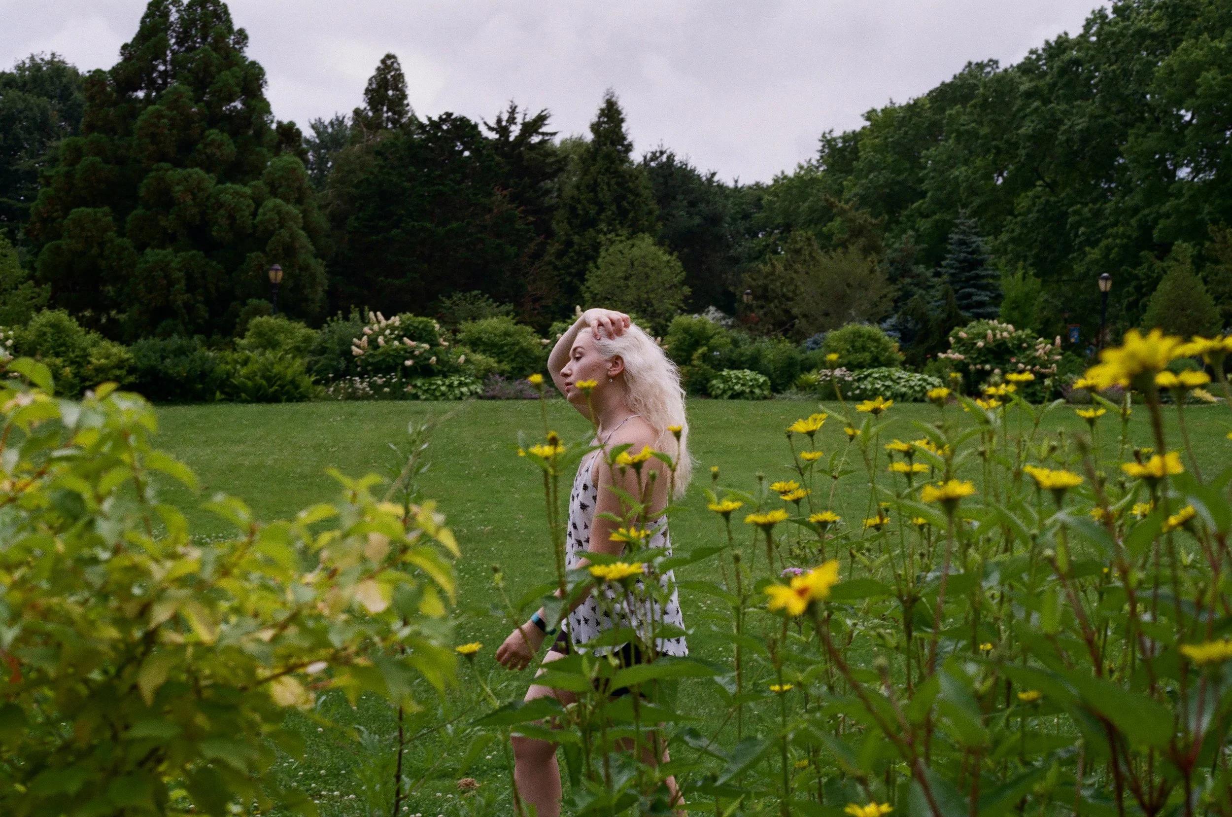A woman with long blonde hair standing among yellow flowers in a park with green trees and bushes in the background.