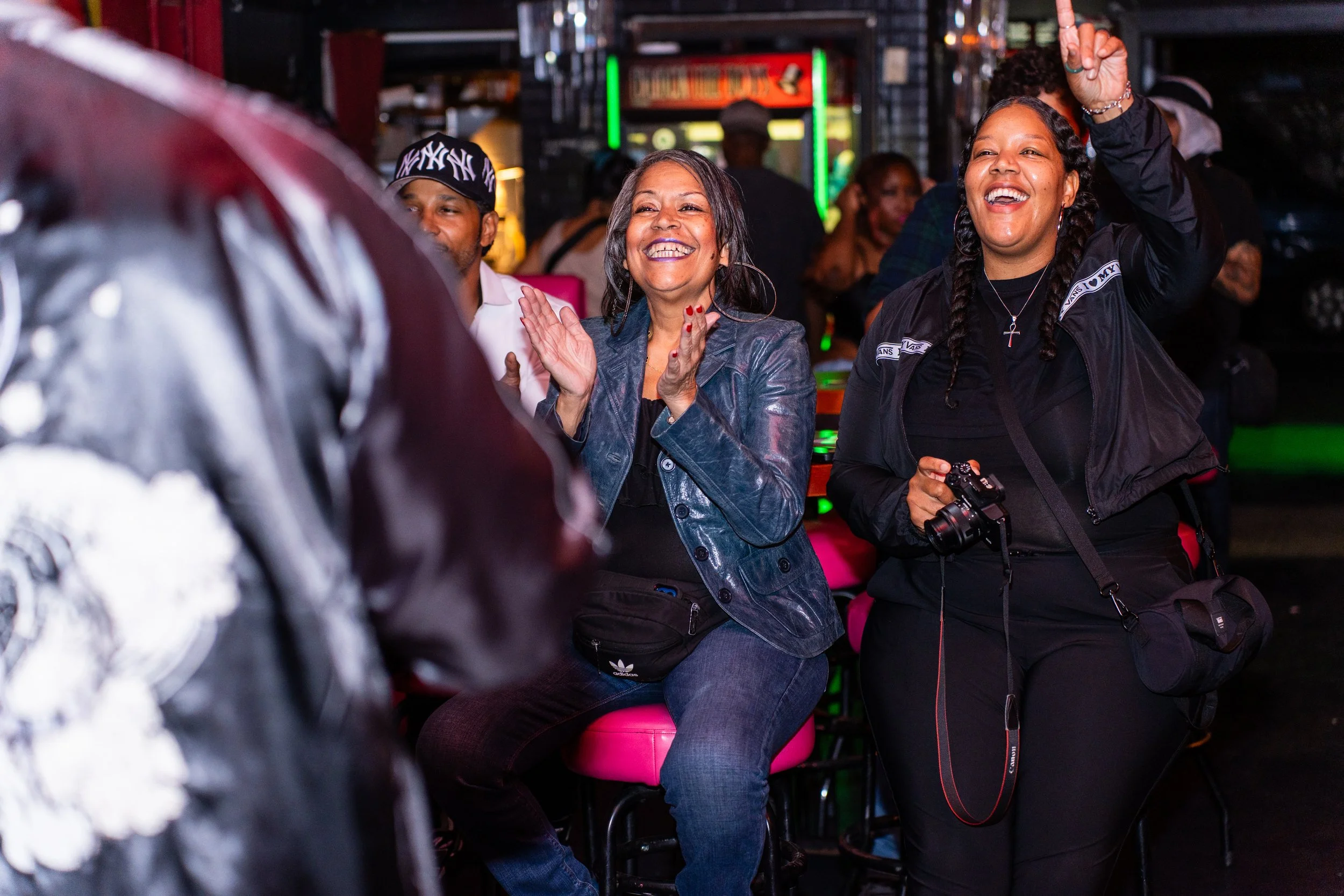 Two women sitting and clapping at an indoor event, others in the background, man with a camera, lively atmosphere, colorful lighting.