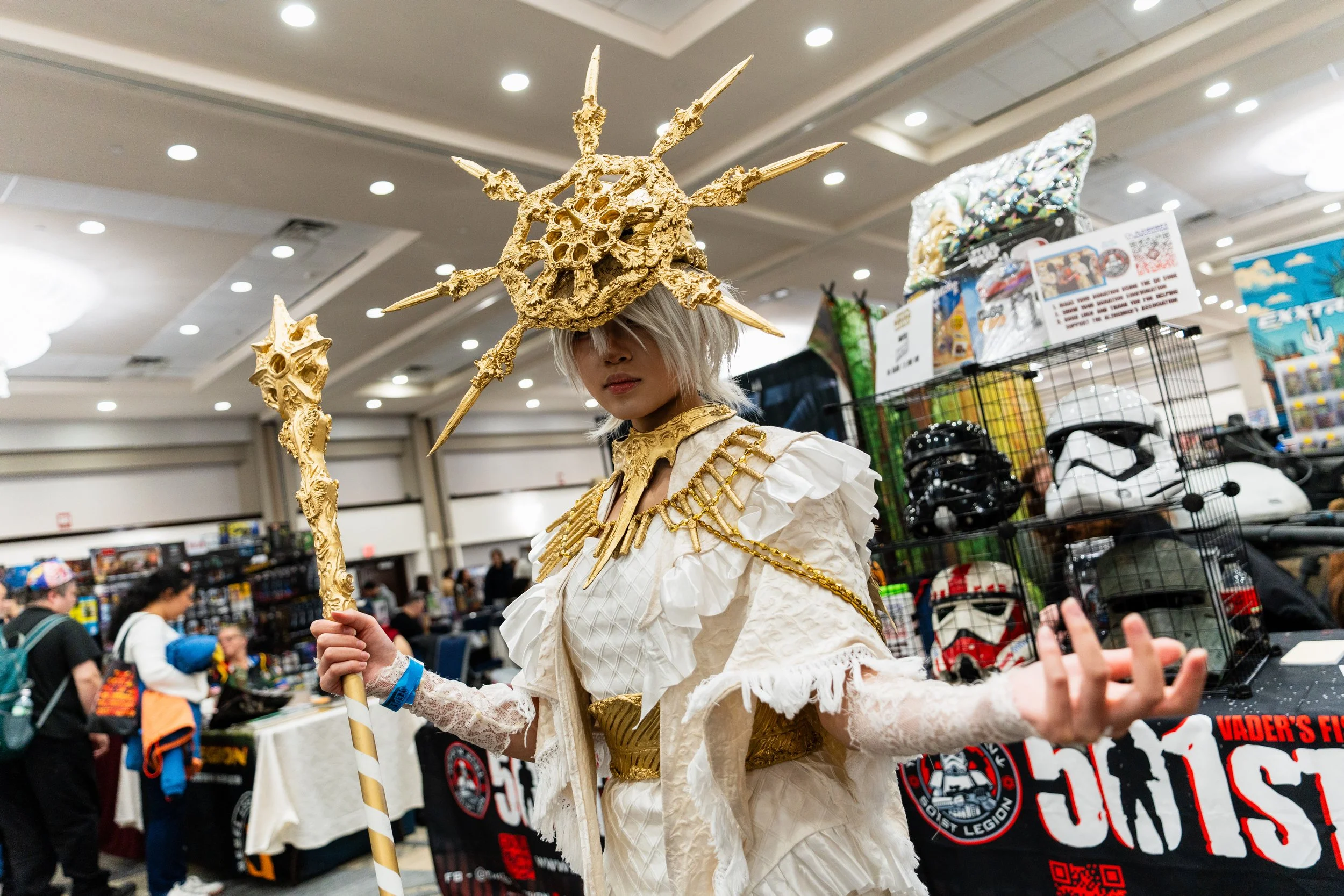 Person dressed in elaborate white and gold costume with a large, ornate gold headpiece resembling spikes or rays, holding a staff, at a convention or event.