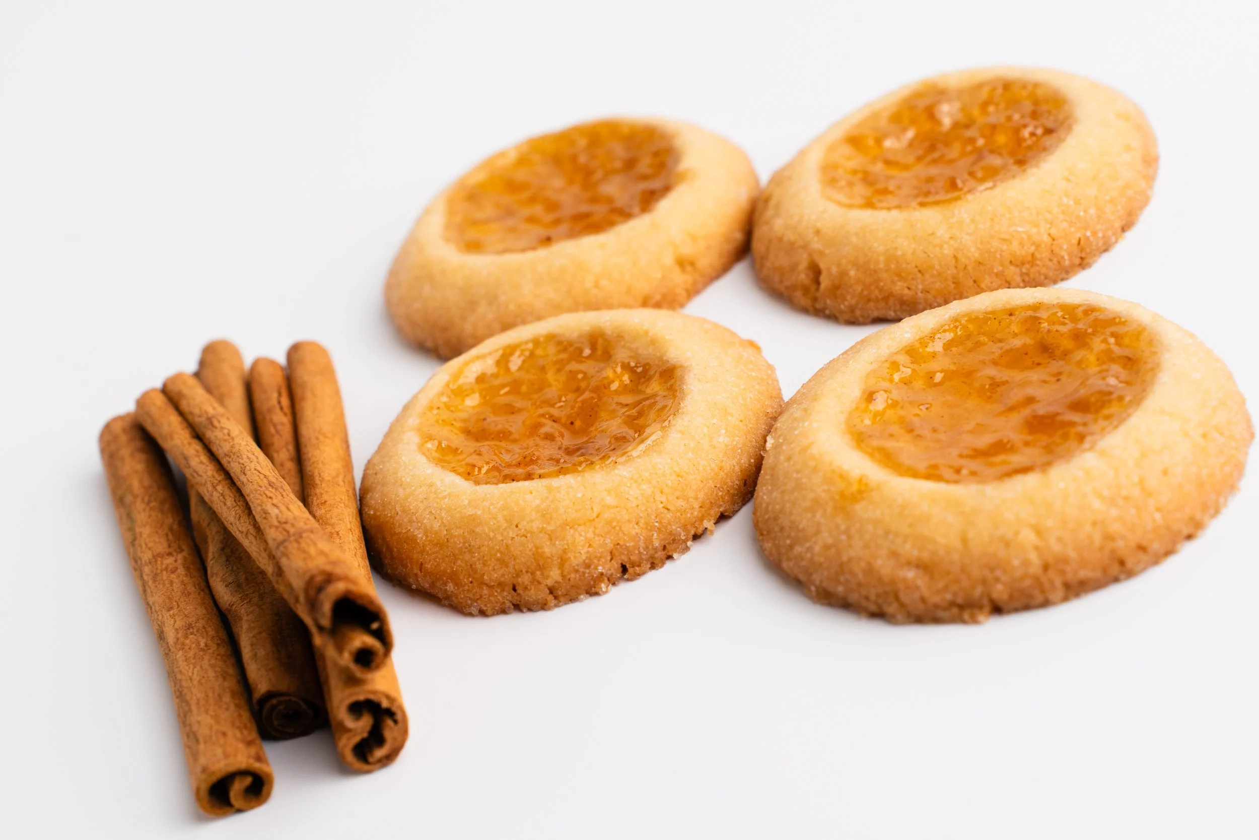Four thumbprint cookies with orange jam filling and a small bunch of cinnamon sticks on a white background.