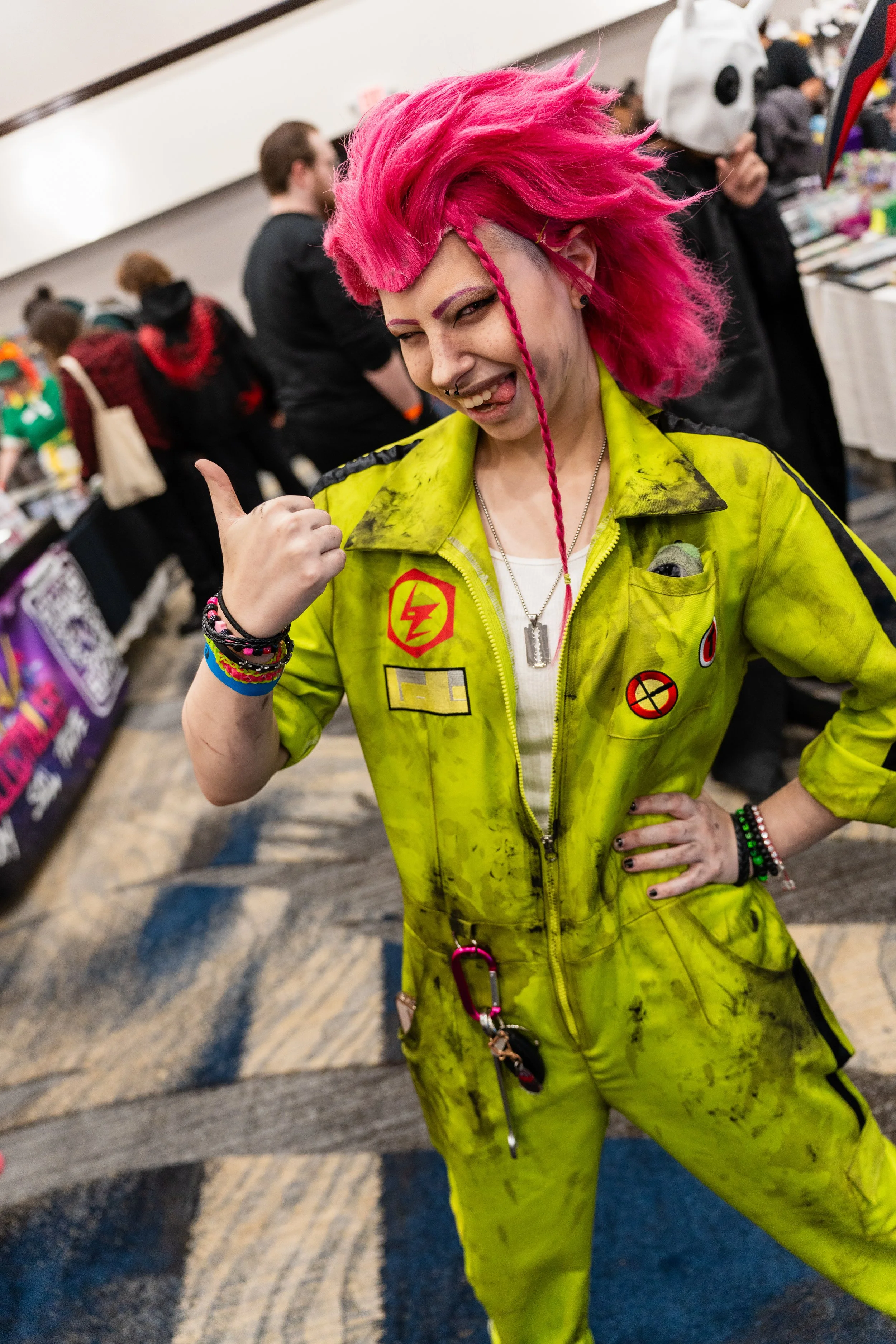A woman with bright pink hair styled in a mohawk and braid, wearing a yellow jumpsuit with patches, giving a thumbs-up and smiling at the camera.