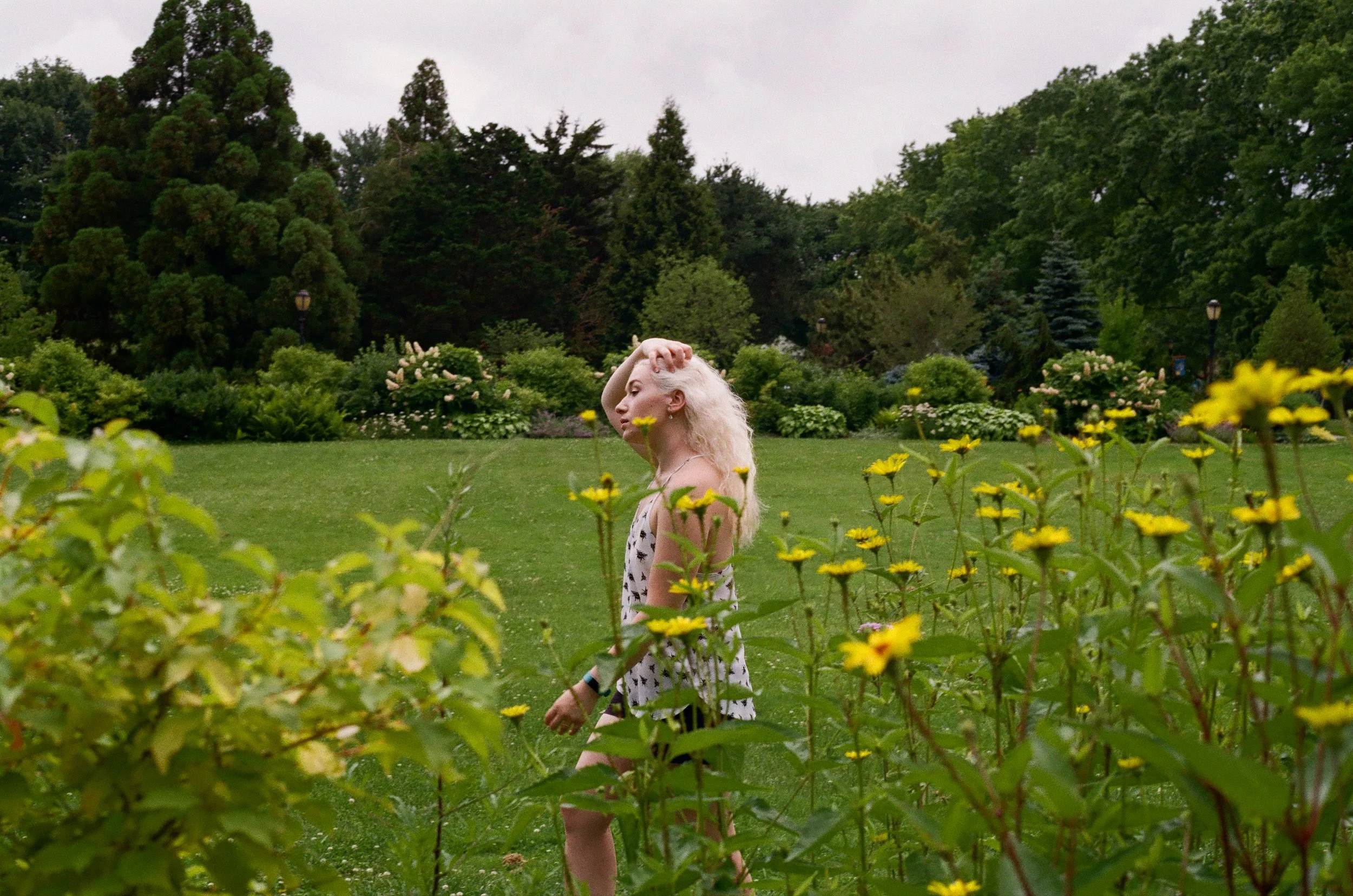 A young blonde woman in a white dress with black polka dots standing in a garden with yellow flowers and greenery, with trees and bushes in the background.