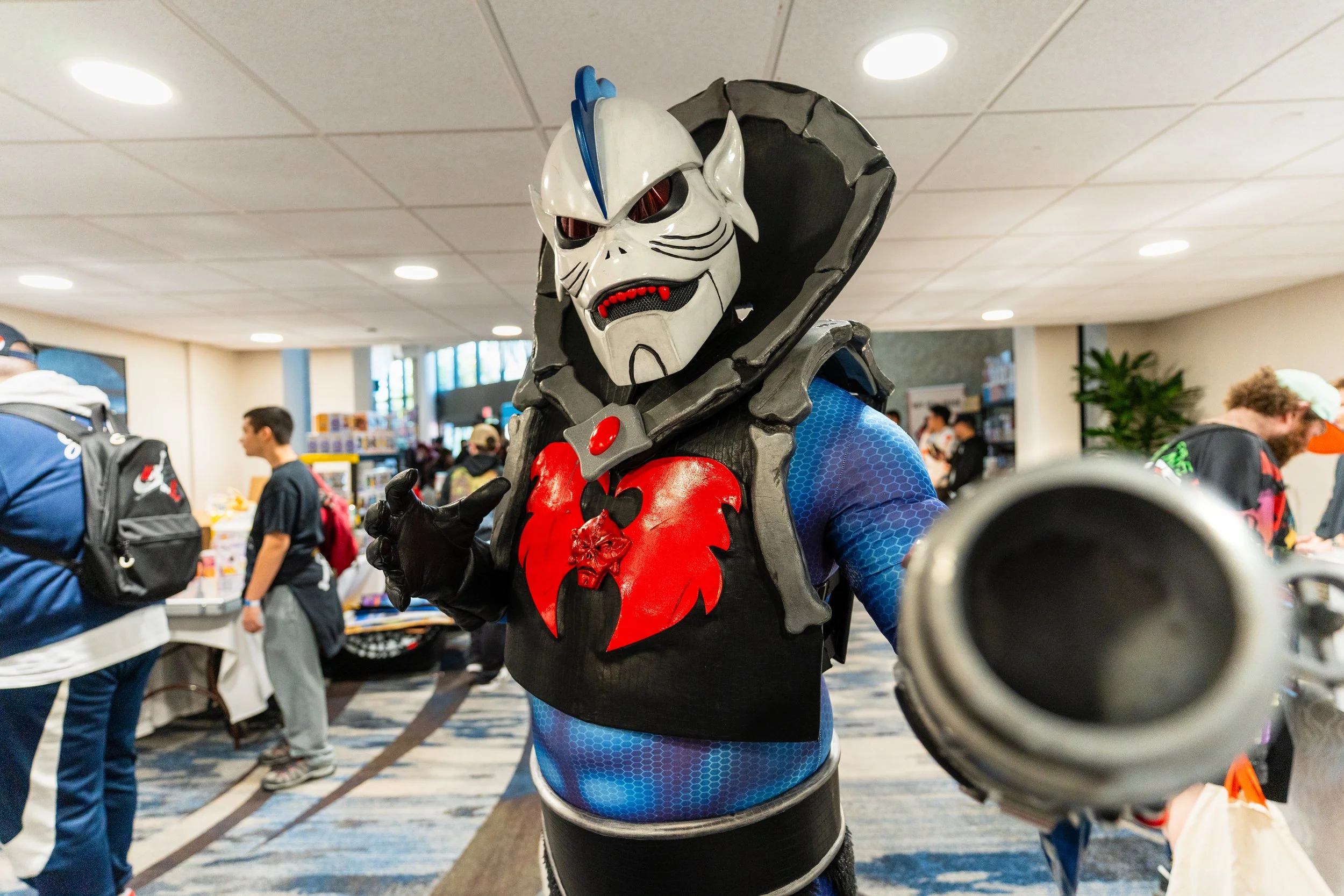 A person in a detailed skeleton warrior costume with a helmet, a black cloak with a red symbol, and blue sleeves, pointing a large prop gun toward the camera at a crowded indoor event.