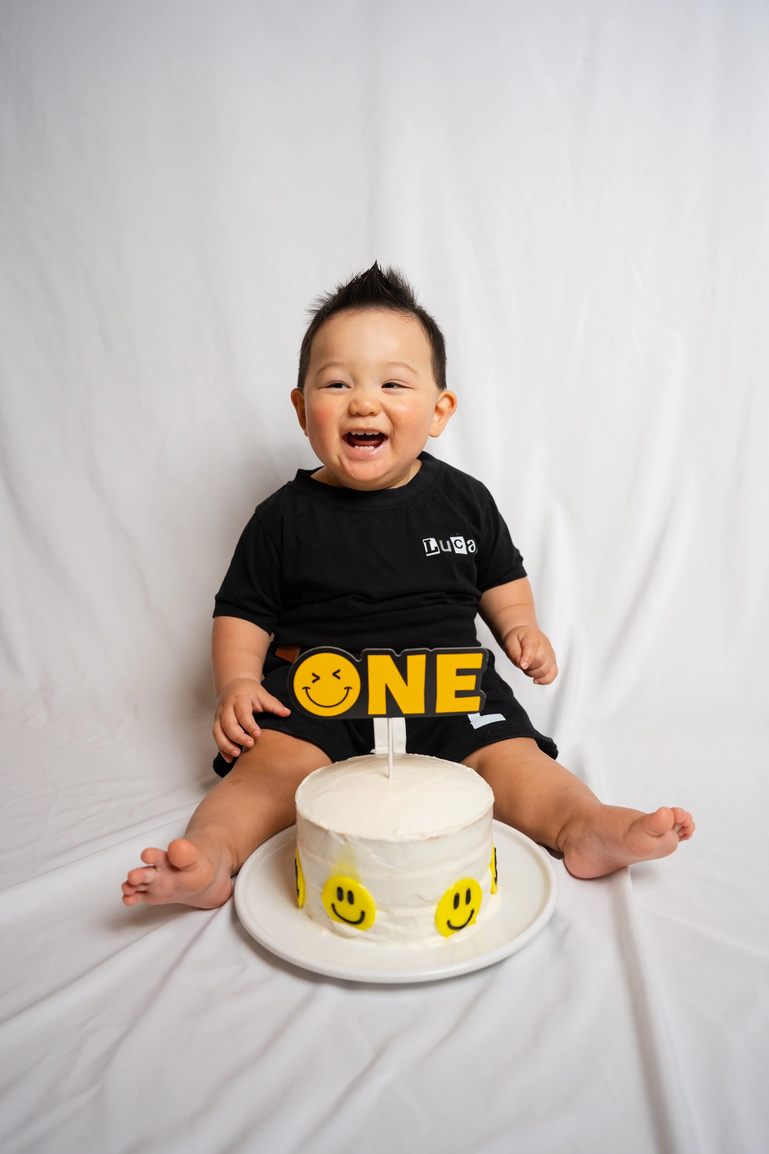 Smiling baby boy sitting in front of a birthday cake with a cake topper that says "ONE". The cake and baby are on a white background.