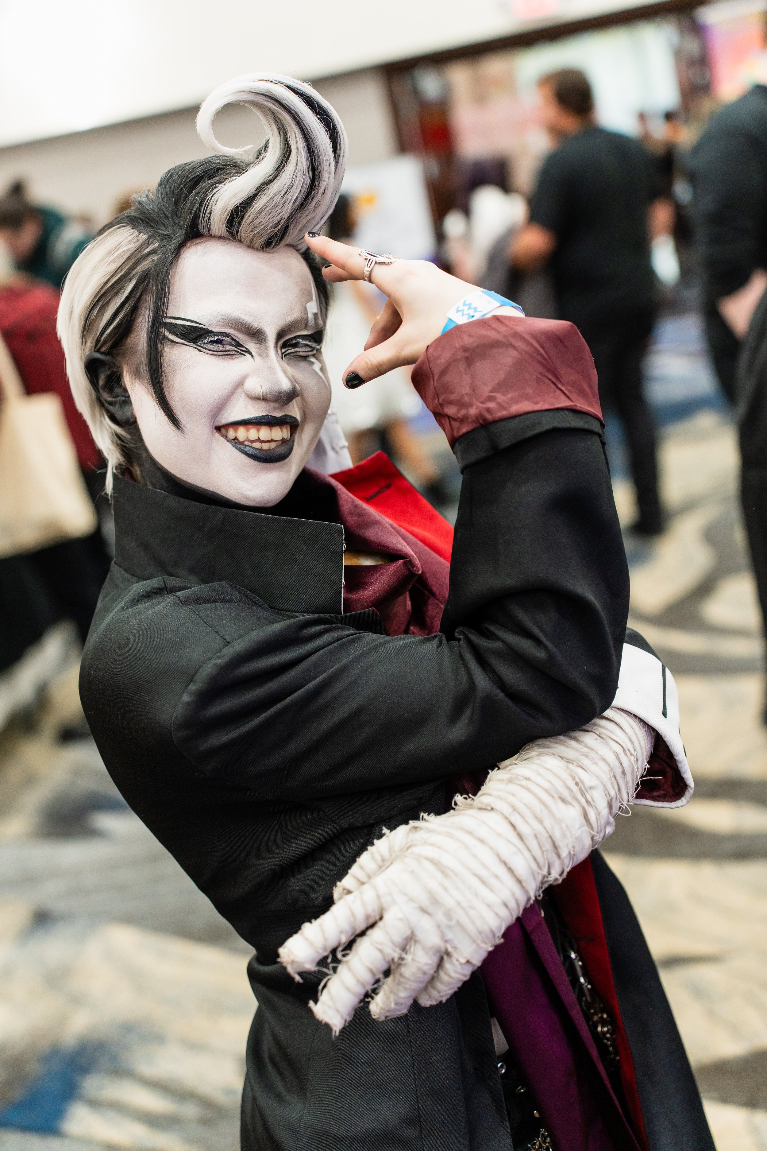 Person dressed as a gothic character with black and white makeup, black lipstick, and a styled black and white hairdo, smiling and striking a pose at a convention or event.