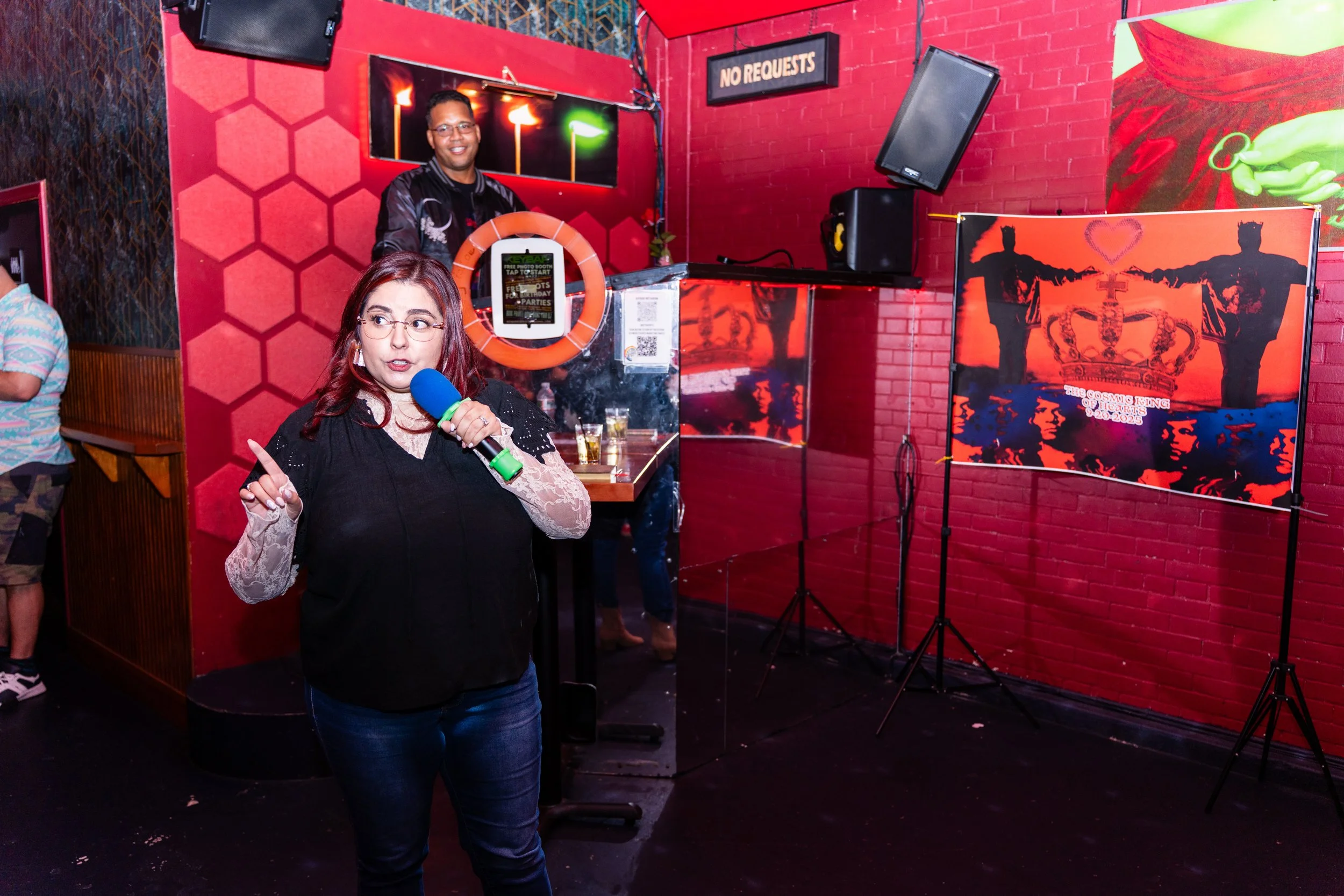A woman holding a microphone and speaking in front of a red brick wall with artworks and speakers, with a man smiling and standing behind a DJ booth in the background.