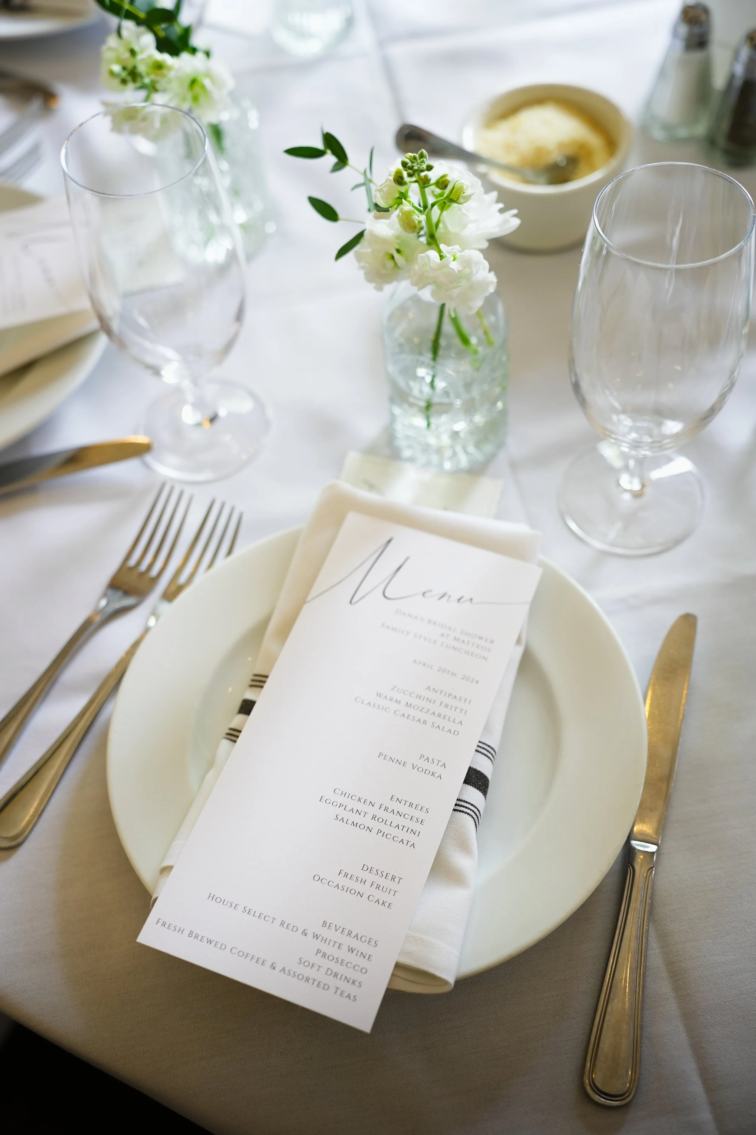 Elegant table setting with a white plate, gold utensils, a printed menu on a napkin, a small floral arrangement in a glass jar, and two empty wine glasses.