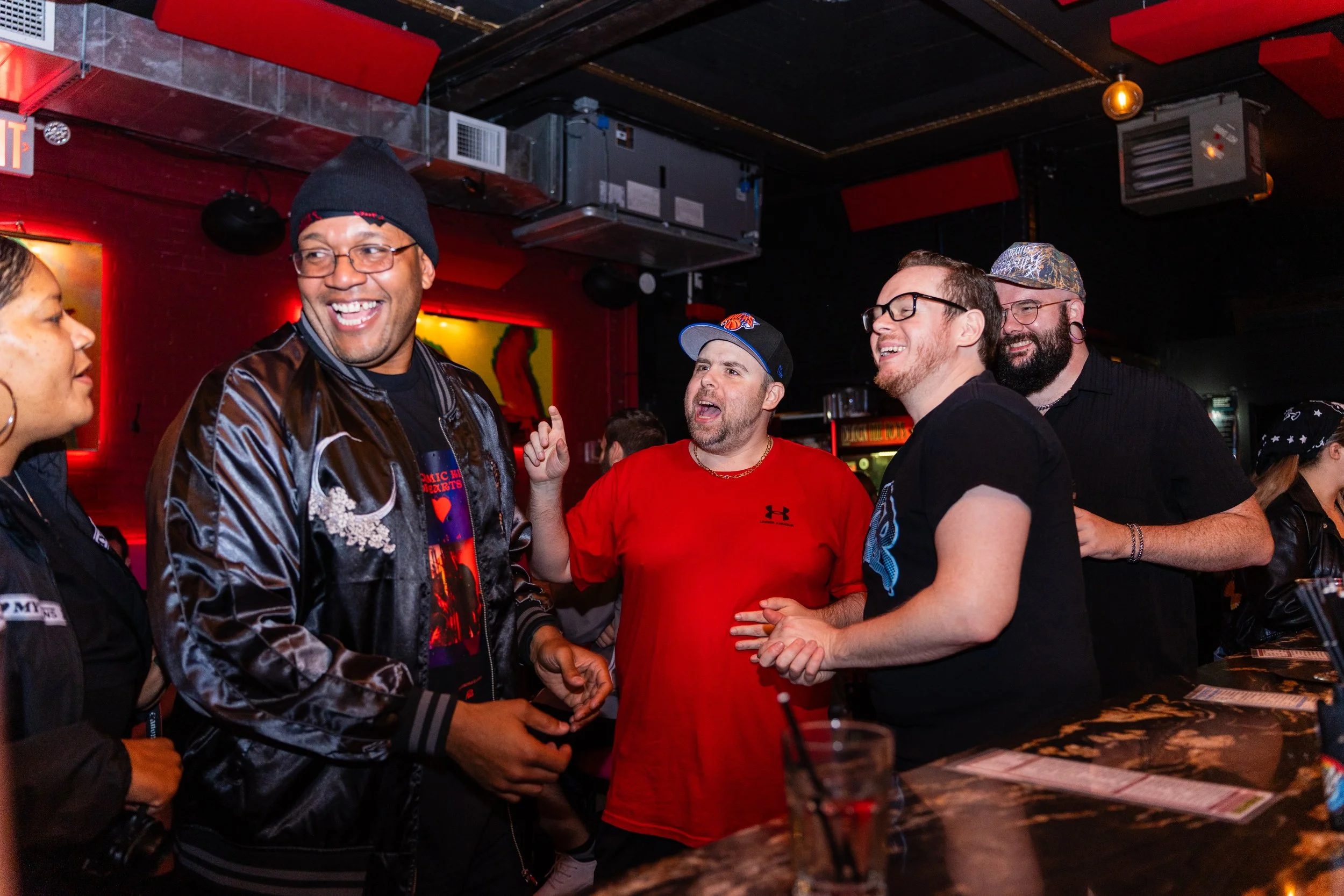 Group of diverse people having a lively conversation and laughing at a bar in a dimly lit nightclub with red walls and artwork in the background.