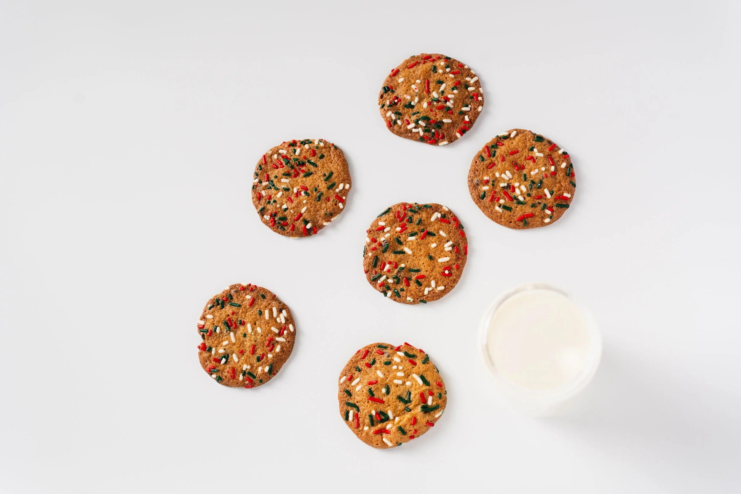 Six chocolate cookies decorated with red, white, green, and gold sprinkles, and a glass of milk on a white surface.