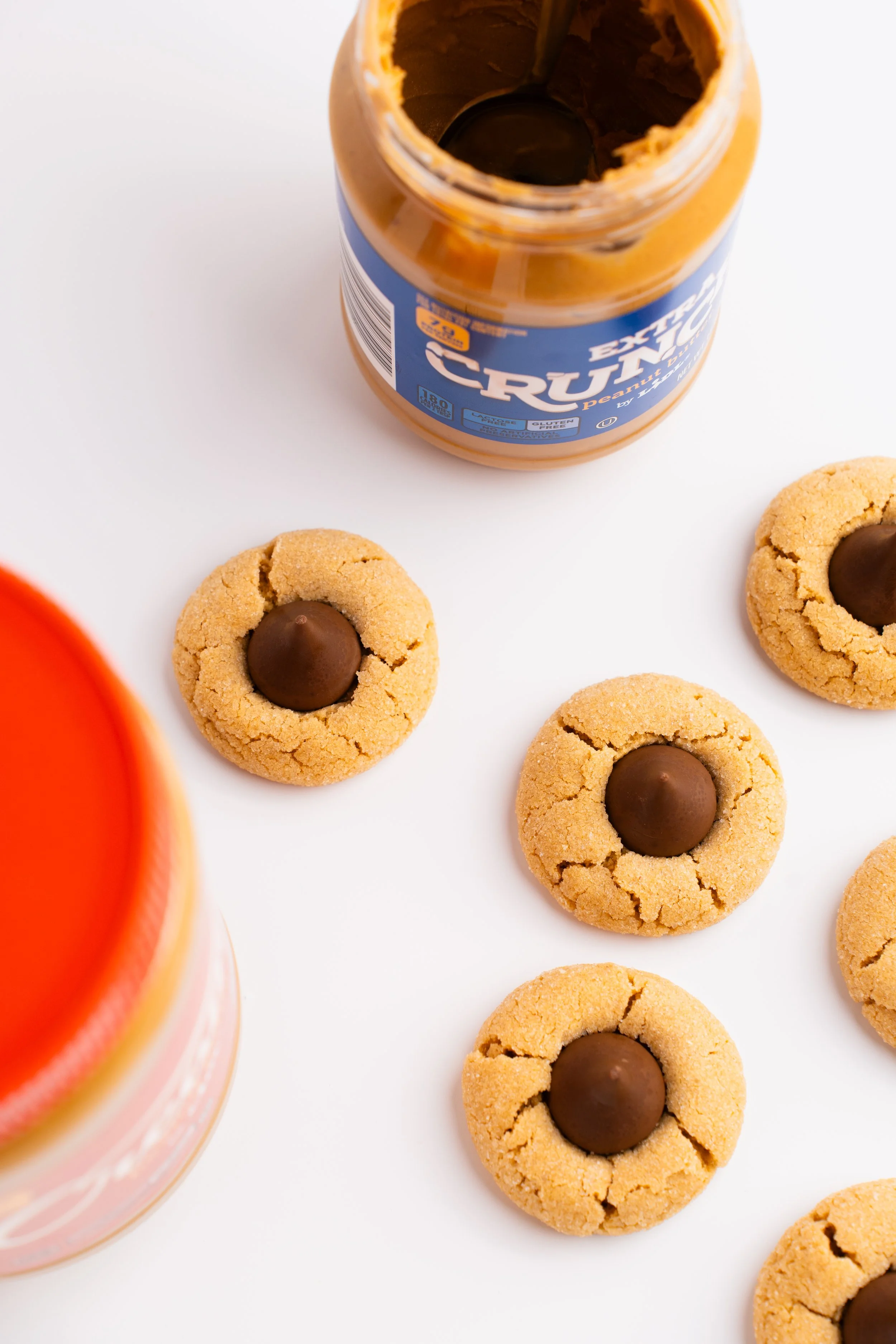 Peanut butter cookies with chocolate center on a white surface with a jar of peanut butter, a container of orange juice, and a jar of jam in the background.