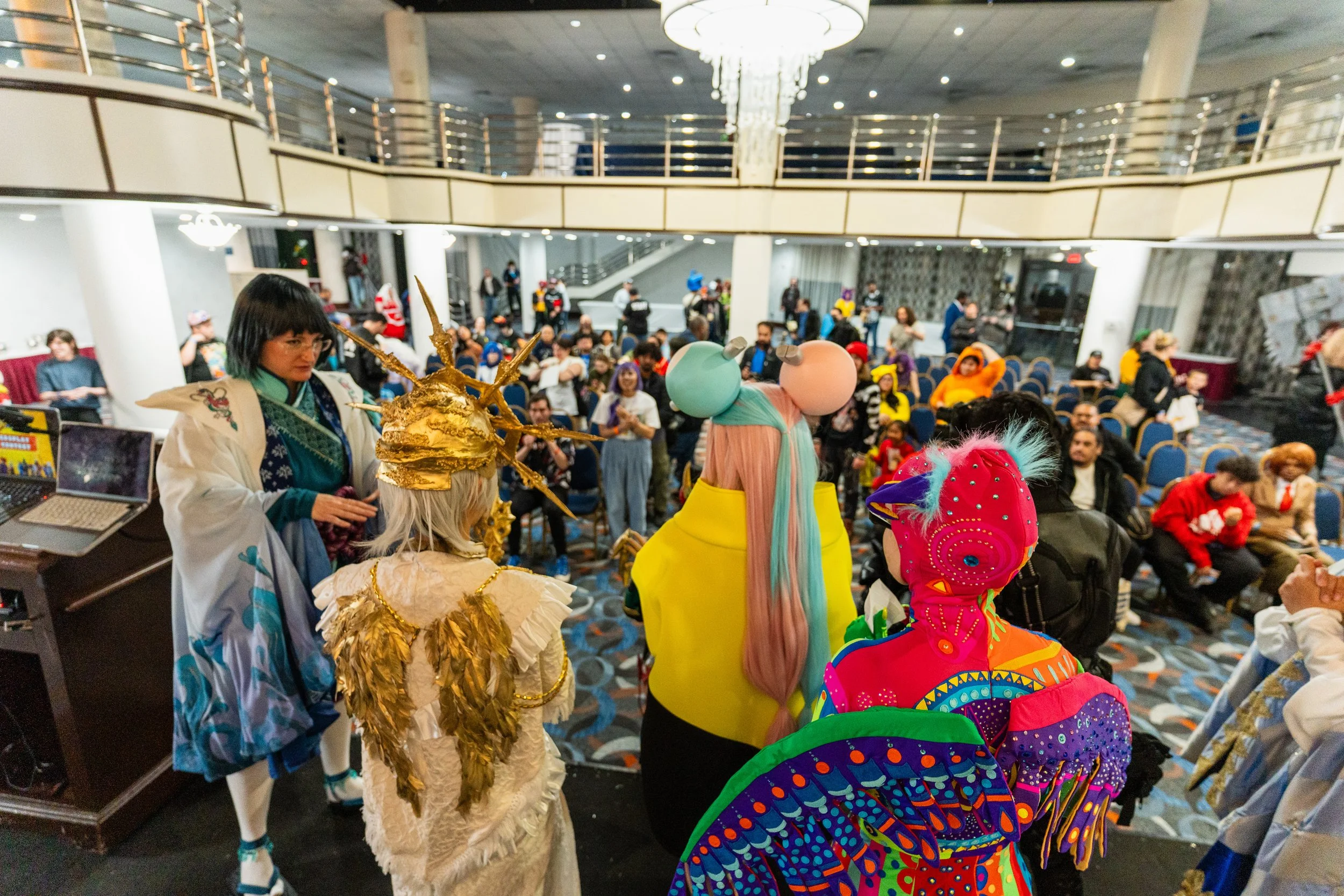 People in colorful costumes at a convention in a large interior space with a second-floor balcony and chandelier.
