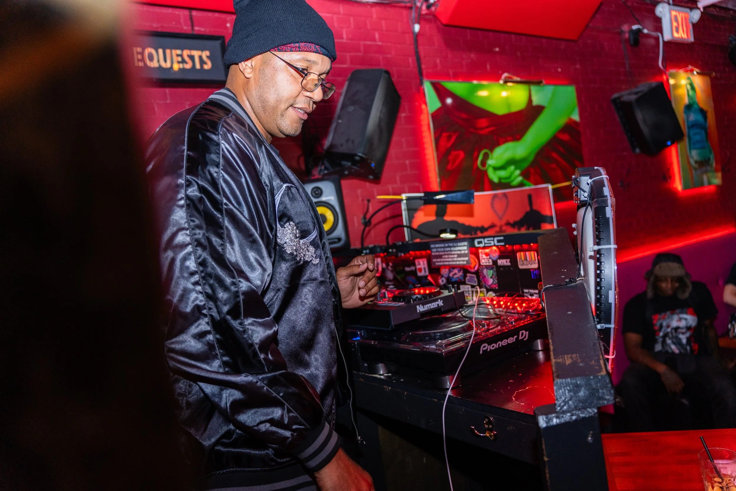 A DJ wearing glasses, a black beanie, and a shiny jacket, playing music at a nightclub with red brick walls and colorful artwork including a painting of a hand holding a green object. There are speakers and mixing equipment on the table.